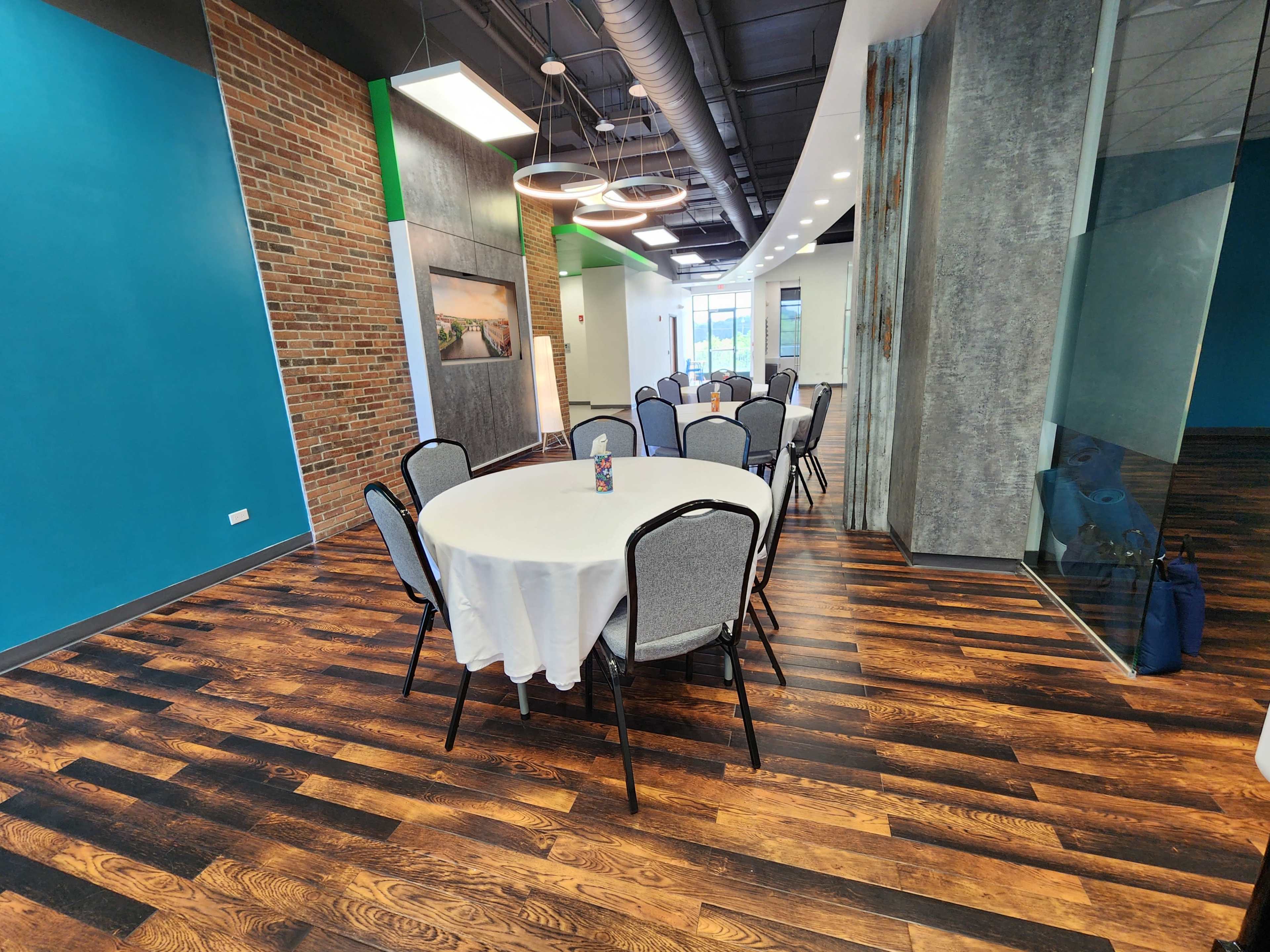 A round table with a white tablecloth is set in a modern room featuring blue and gray walls, wooden flooring, and overhead lighting.