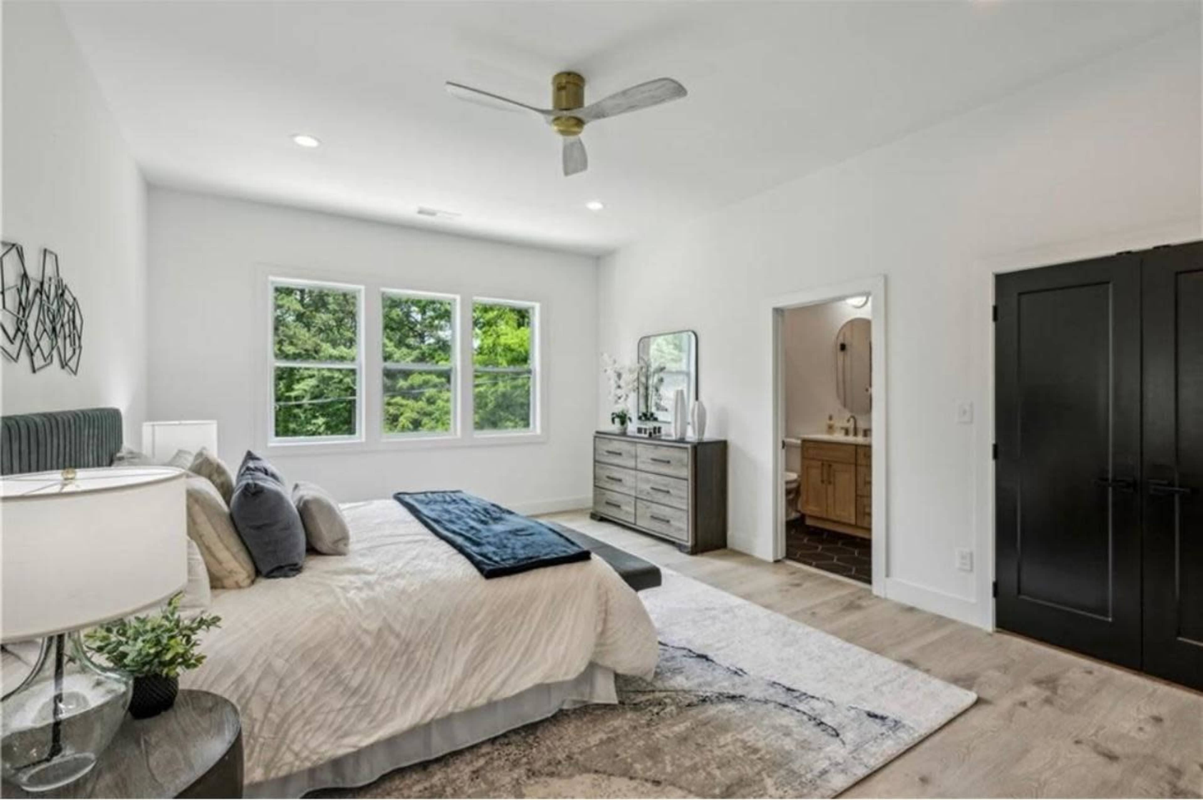 A modern bedroom features a large bed, a dresser, and windows overlooking greenery, with an adjoining bathroom visible in the background.