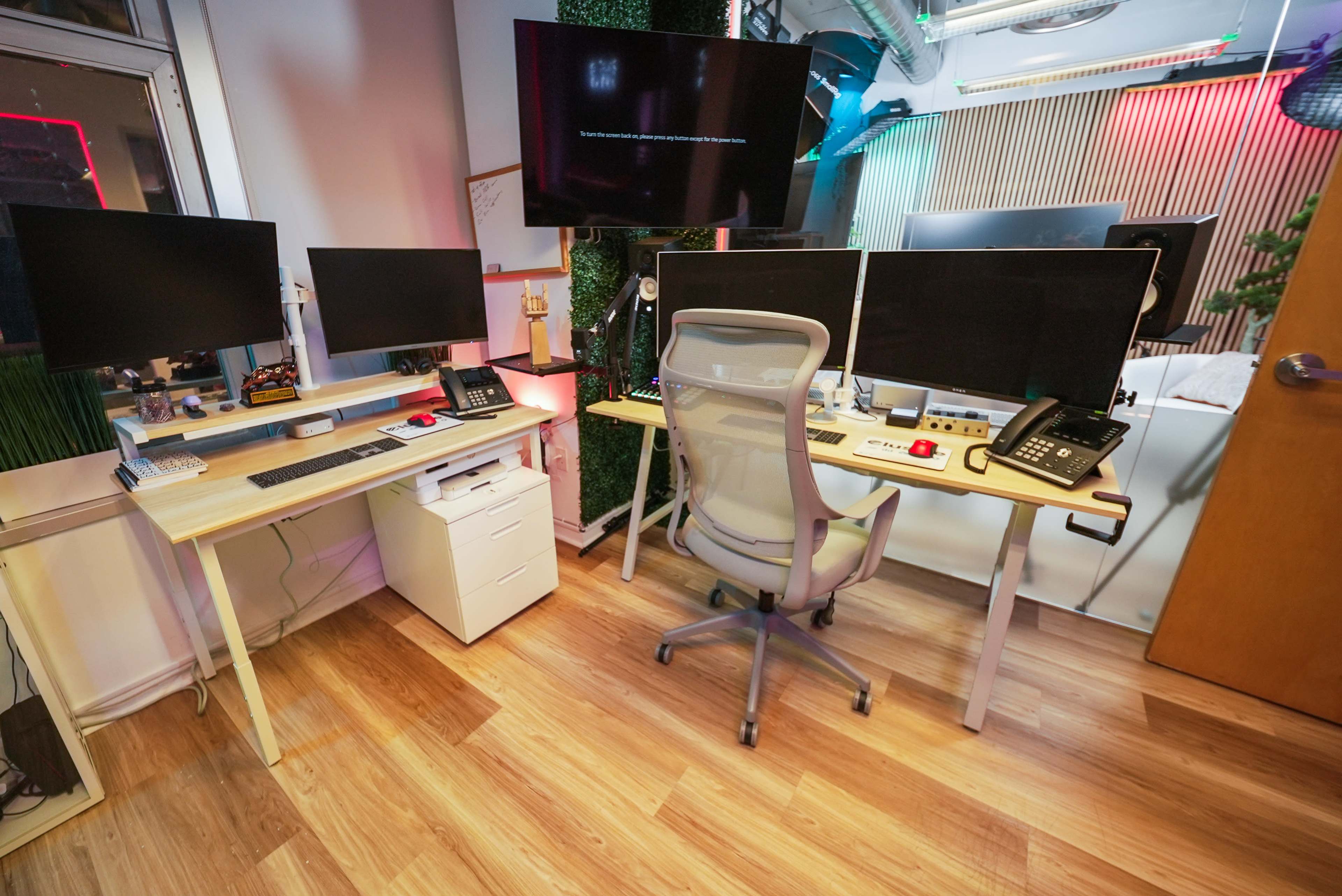 The image shows a modern office space with two desks equipped with multiple monitors, a chair, and various office equipment such as phones and a filing cabinet.