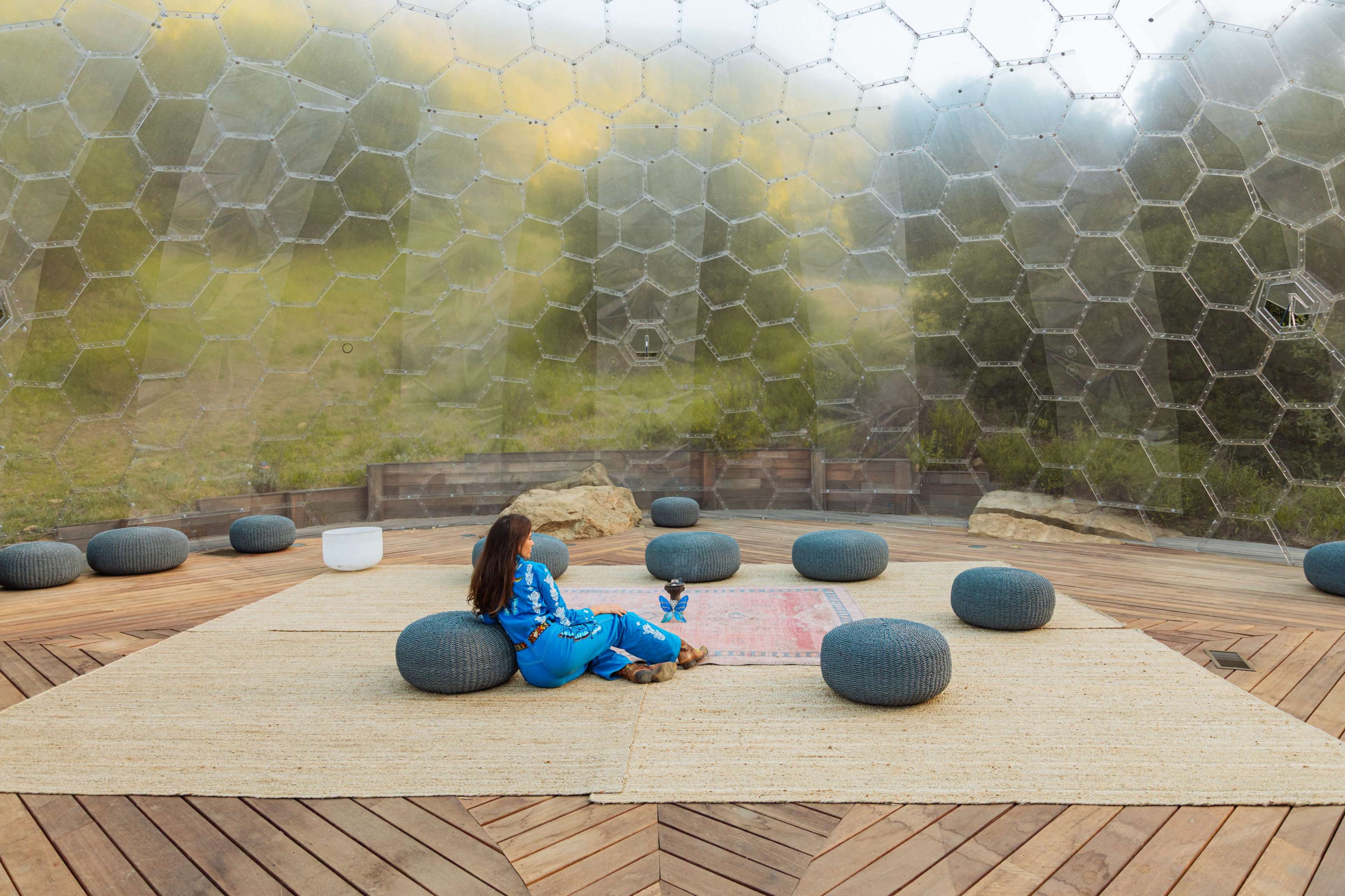 A person seated on a woven rug inside a transparent, hexagonal-shaped structure with circular cushions on a wooden floor.