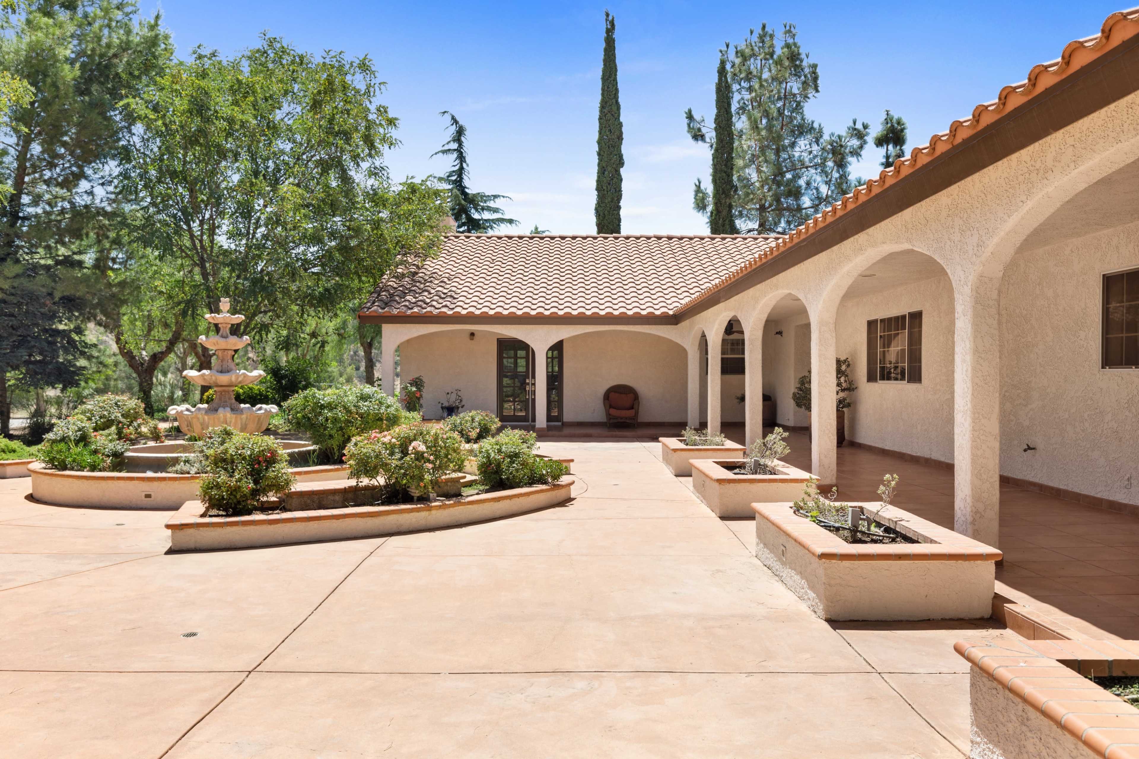 A spacious courtyard features a fountain surrounded by landscaped gardens and an adjoining single-story house with arched entrances.