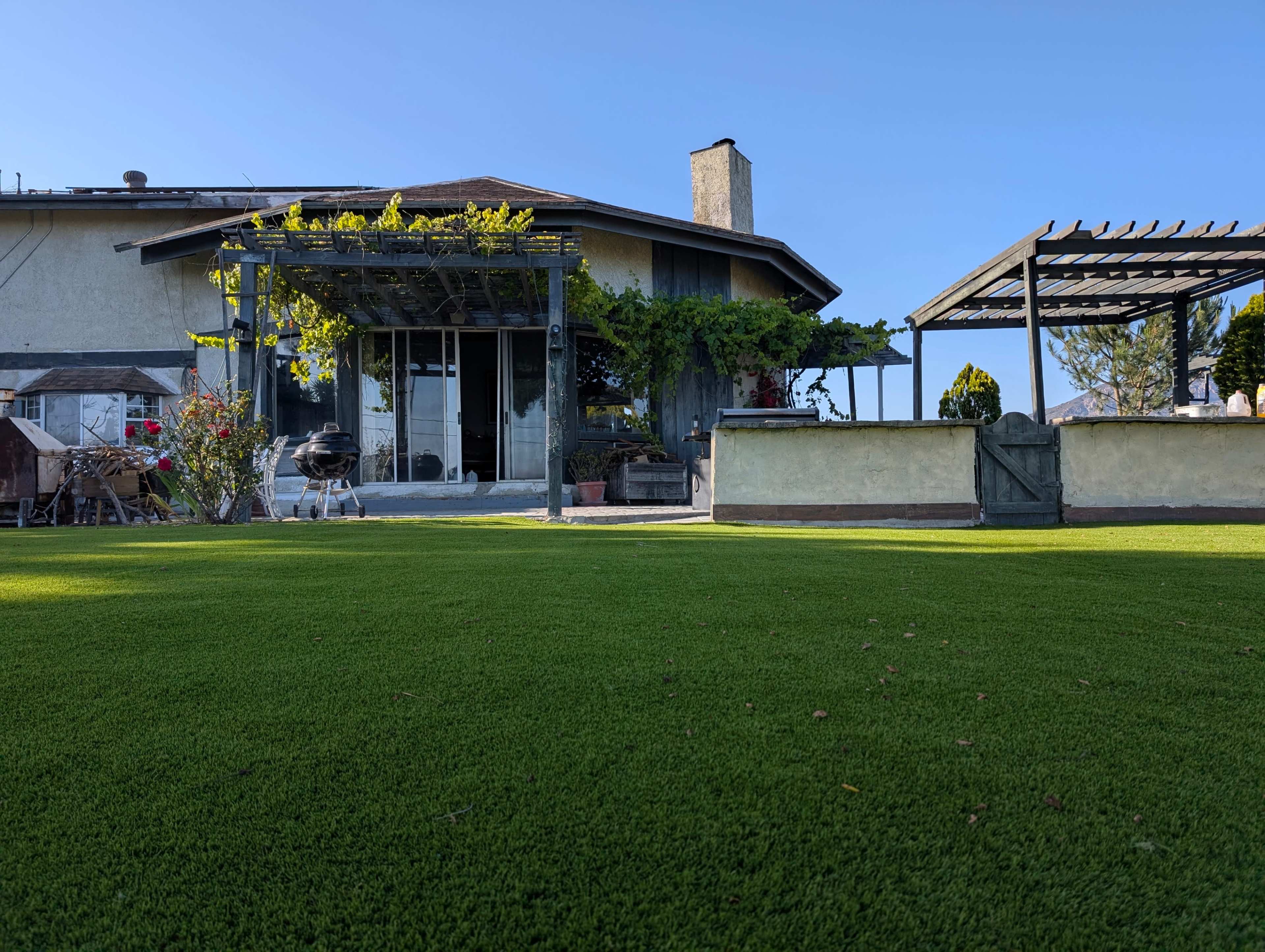 A house with a spacious lawn features a deck with a pergola, potted plants, and a barbecue area.