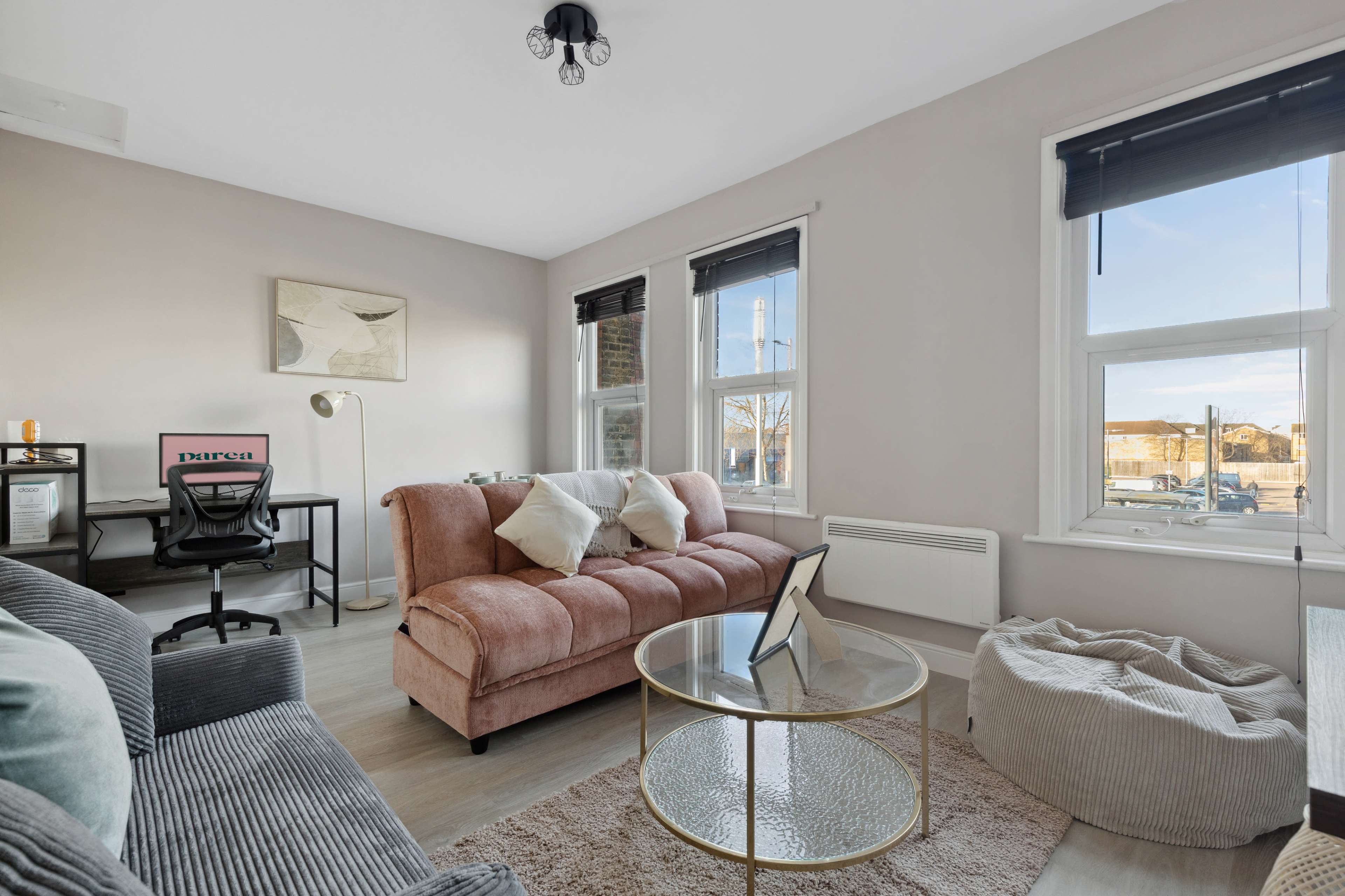 The image shows a living room with a pink sofa, a glass coffee table, a desk with a chair, and large windows providing natural light.