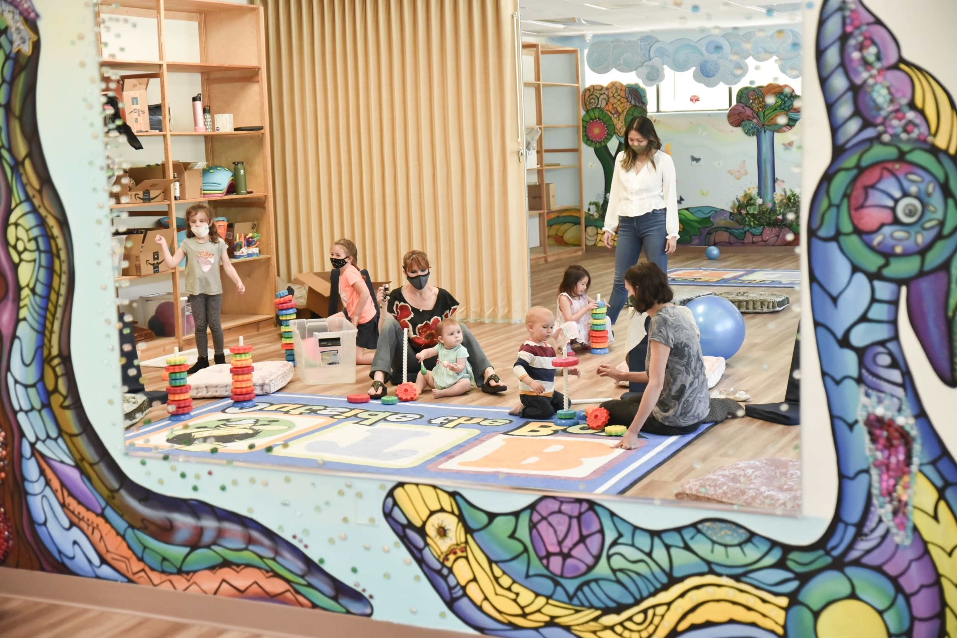 A colorful playroom features children and adults engaged in various activities, including stacking toys and playing on a large mat.