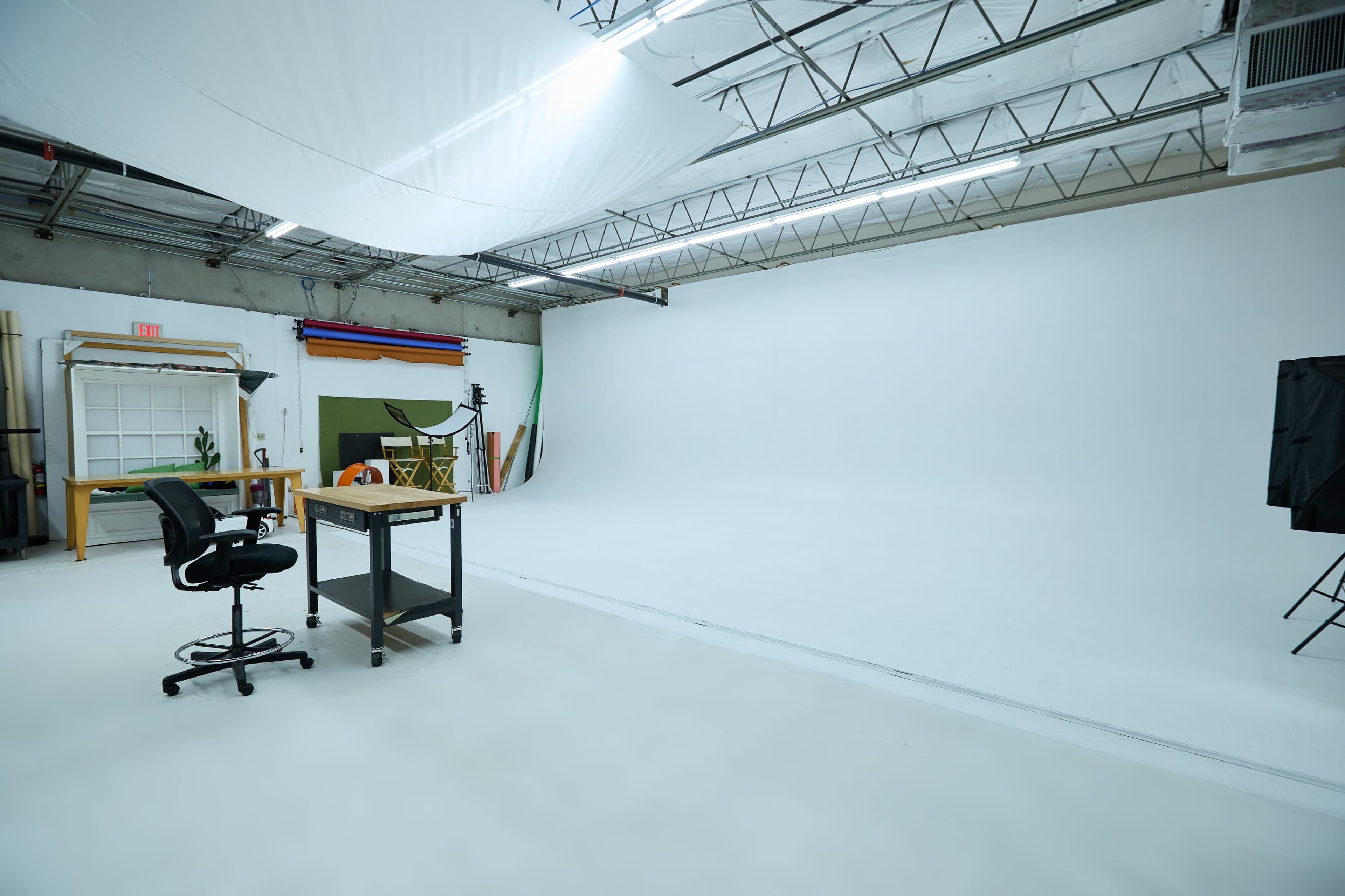 A spacious photography studio features a large white backdrop, a desk with a chair, and various equipment along the walls.