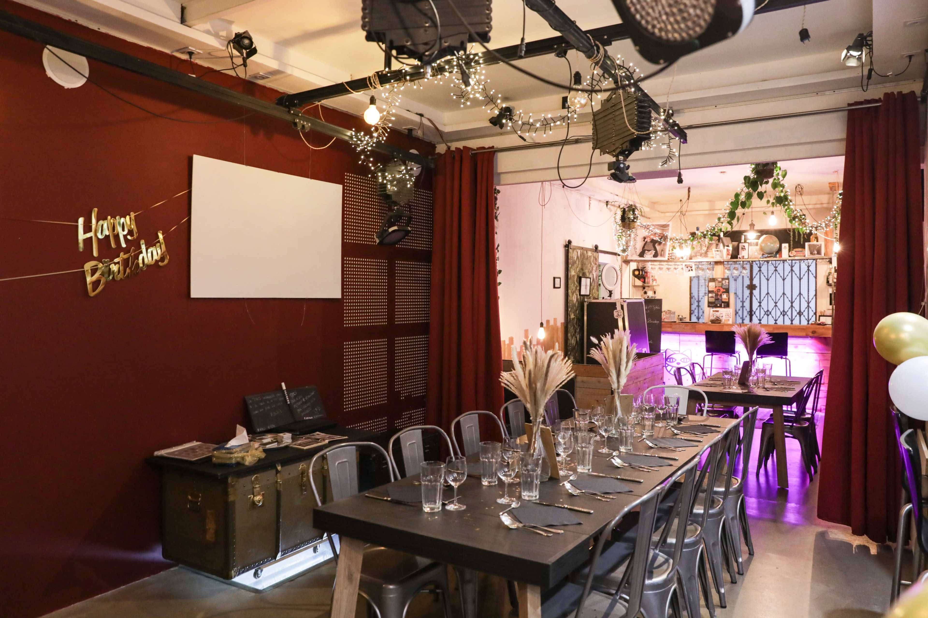 A dining area is set up with a long black table and metal chairs, decorated for a birthday celebration, featuring string lights and a "Happy Birthday" banner on the wall.