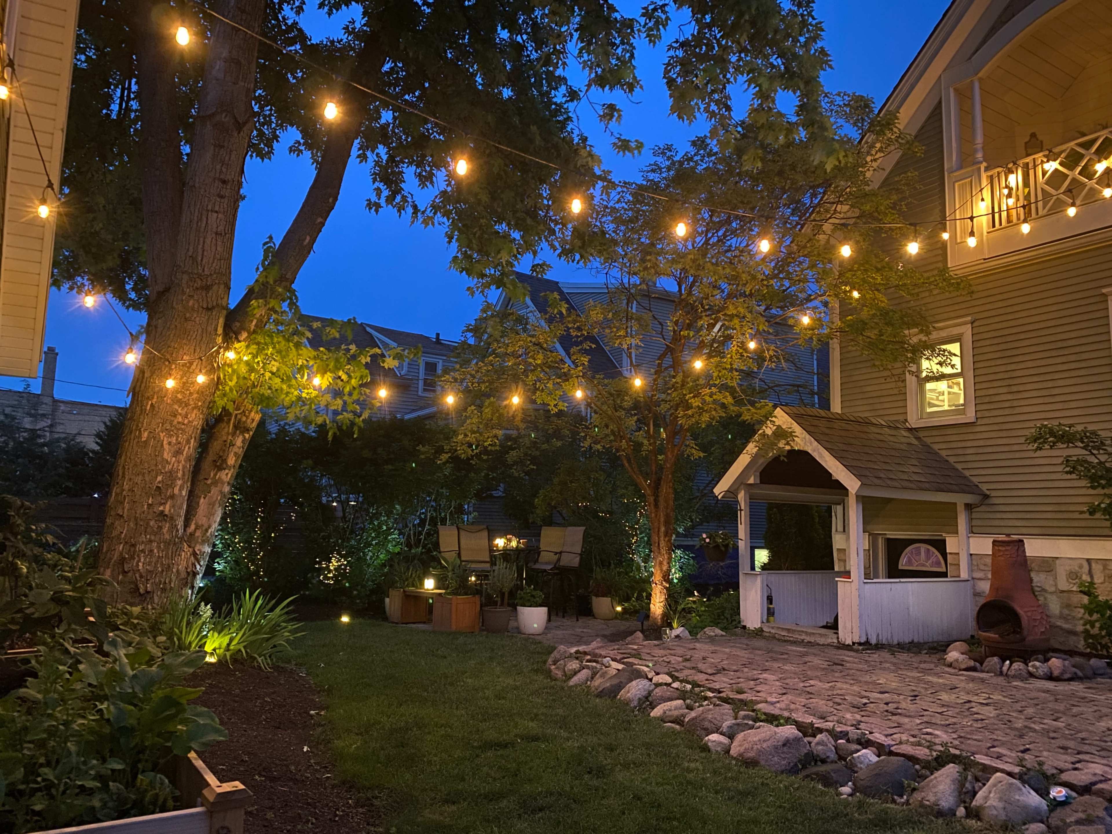The backyard features string lights illuminating a small playhouse, surrounded by greenery and a stone path under a twilight sky.