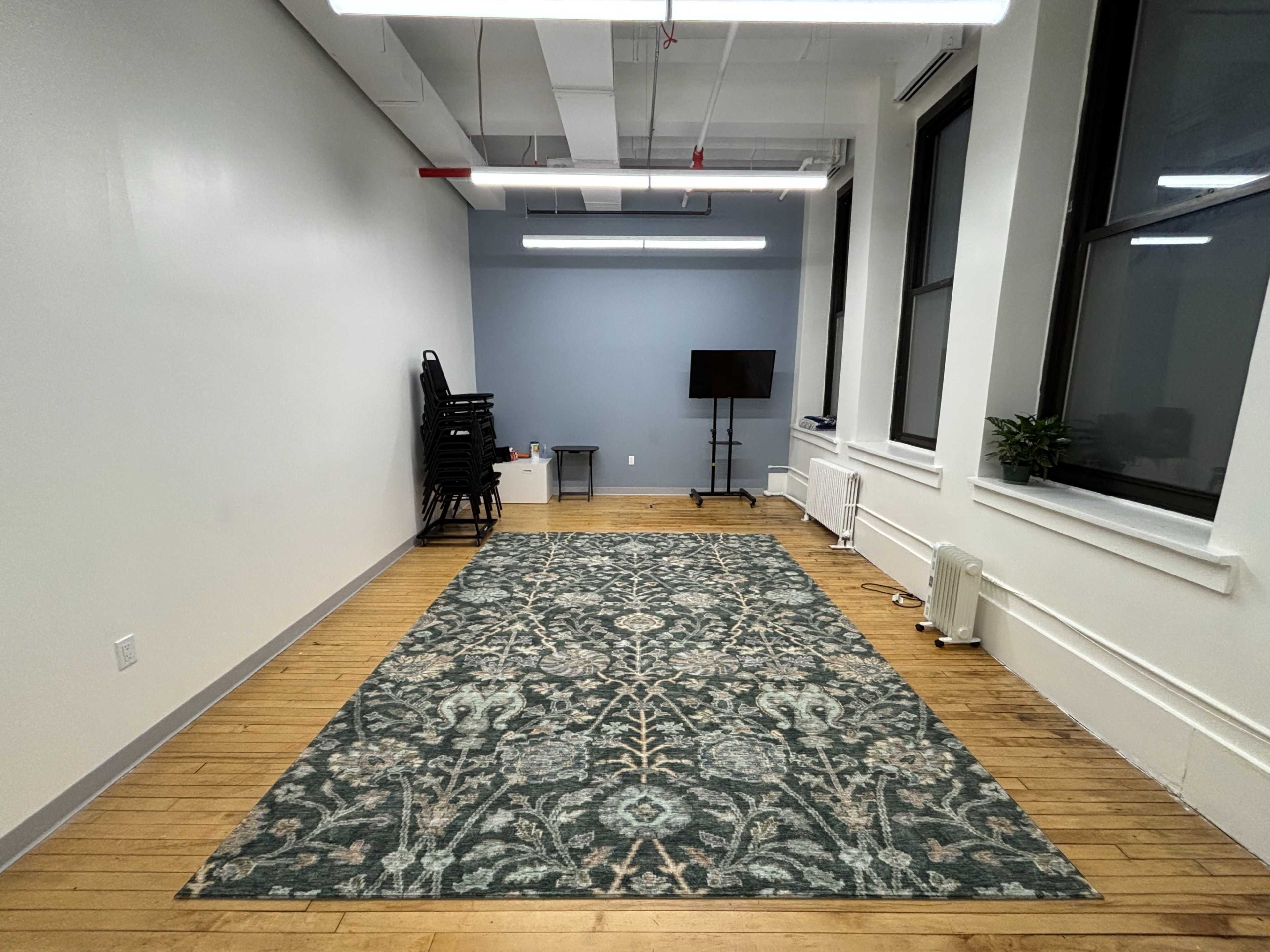 The image shows a large, empty office space with a green patterned rug on the wooden floor, a stack of black chairs against one wall, a table, and a television on a stand.