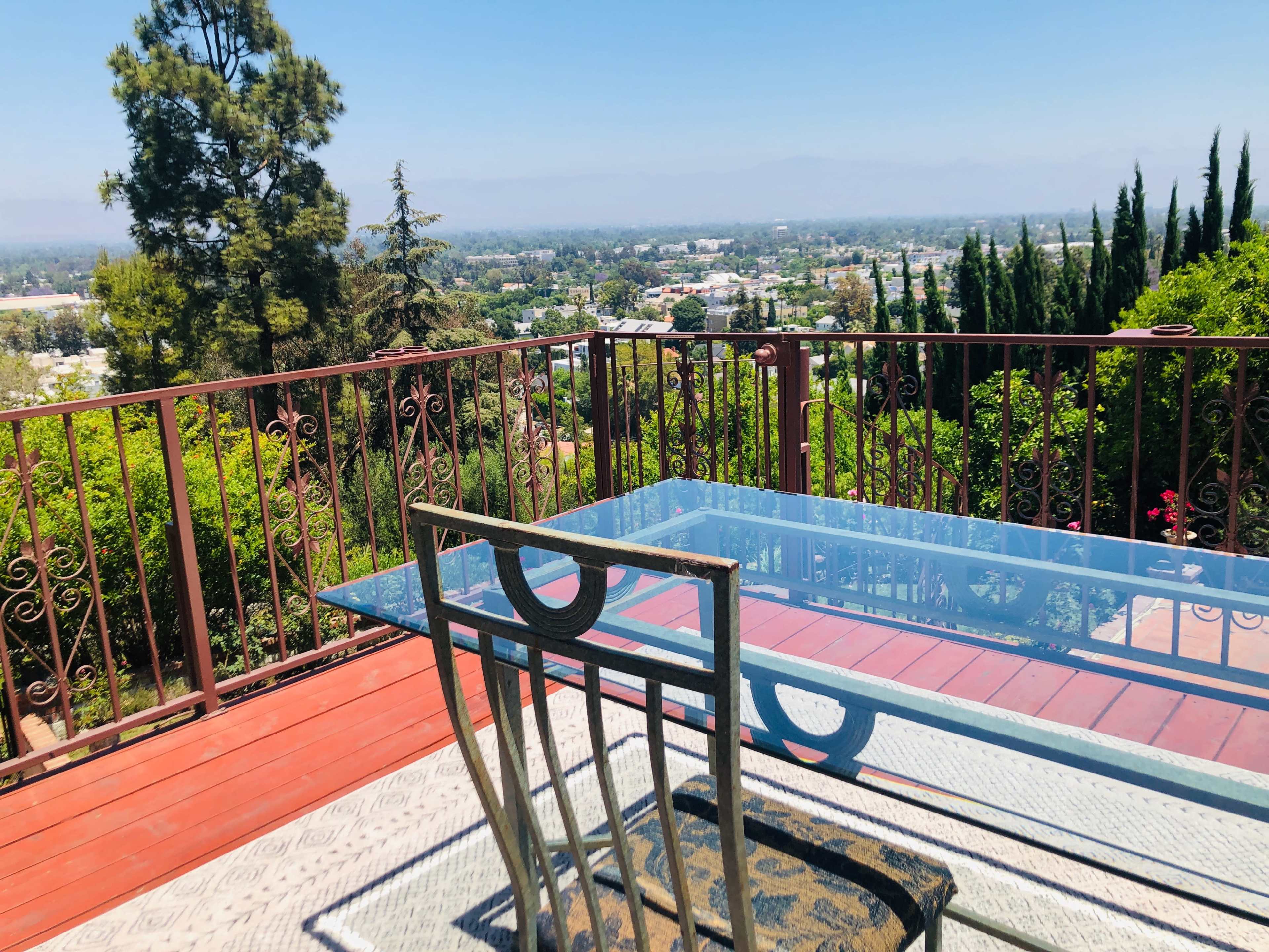A glass table and chair are positioned on a red wooden deck overlooking a green hillside and distant valley.