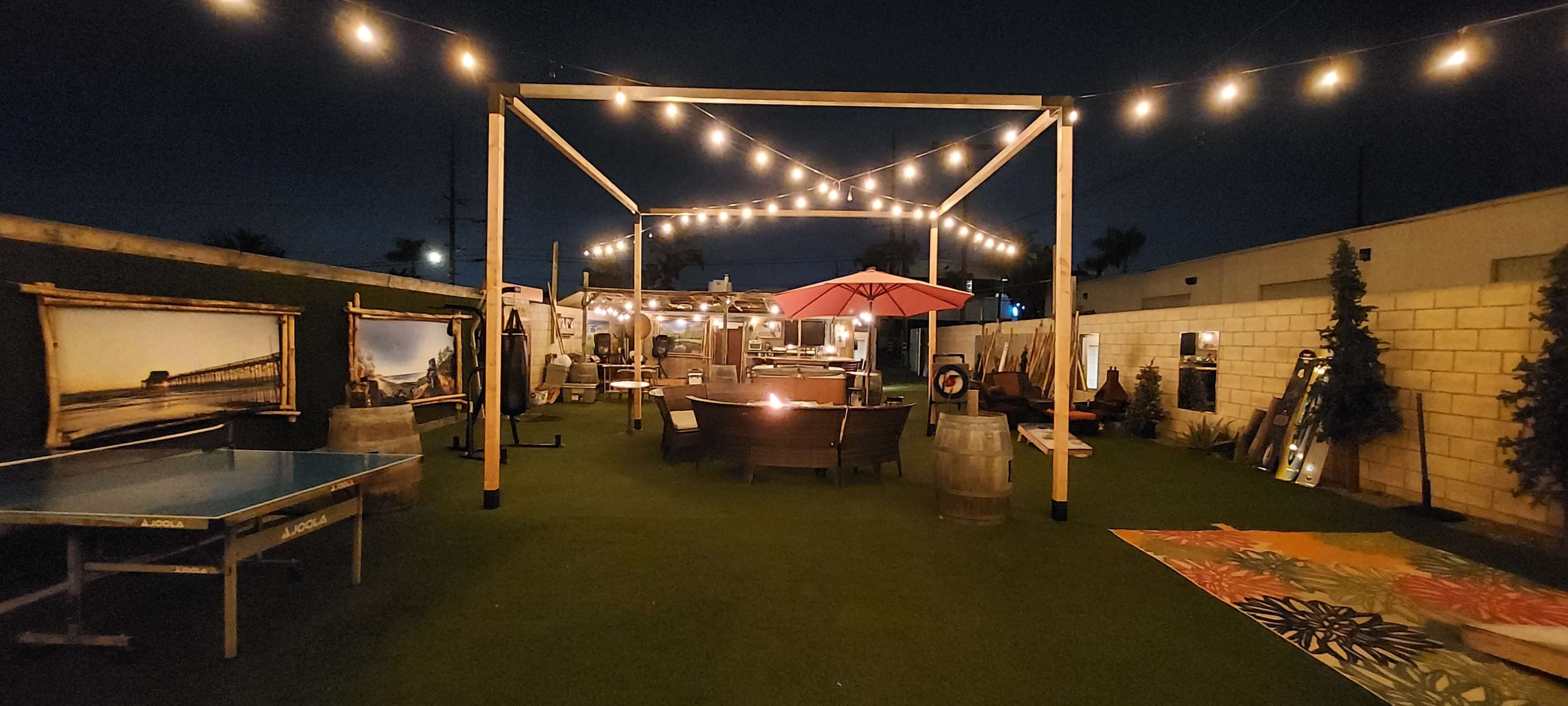 A dimly lit outdoor patio area with string lights, a ping pong table, seating arrangements, and a large umbrella.