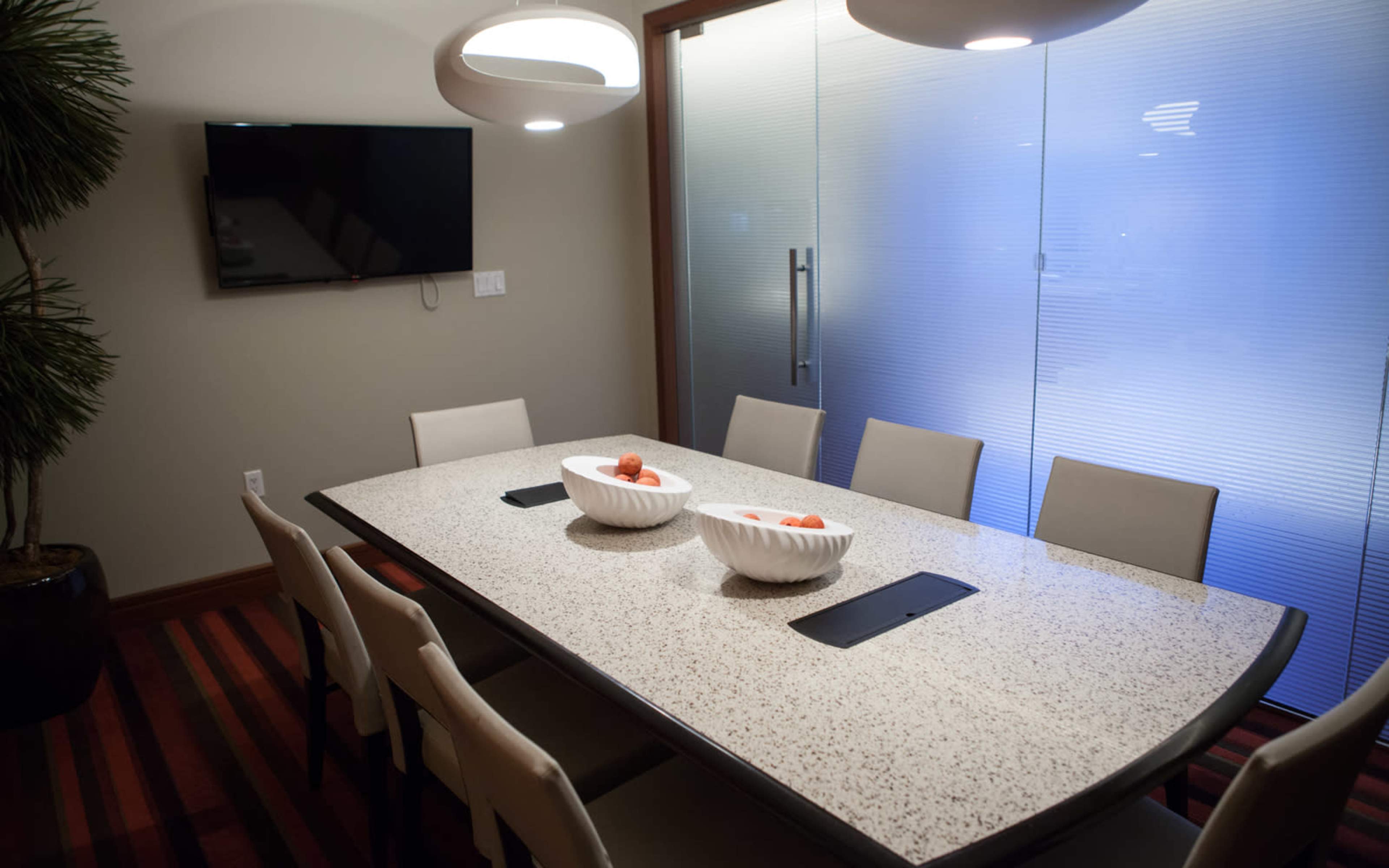 A dining area features a long table with two white bowls of fruit, surrounded by eight chairs, and a television mounted on the wall nearby.