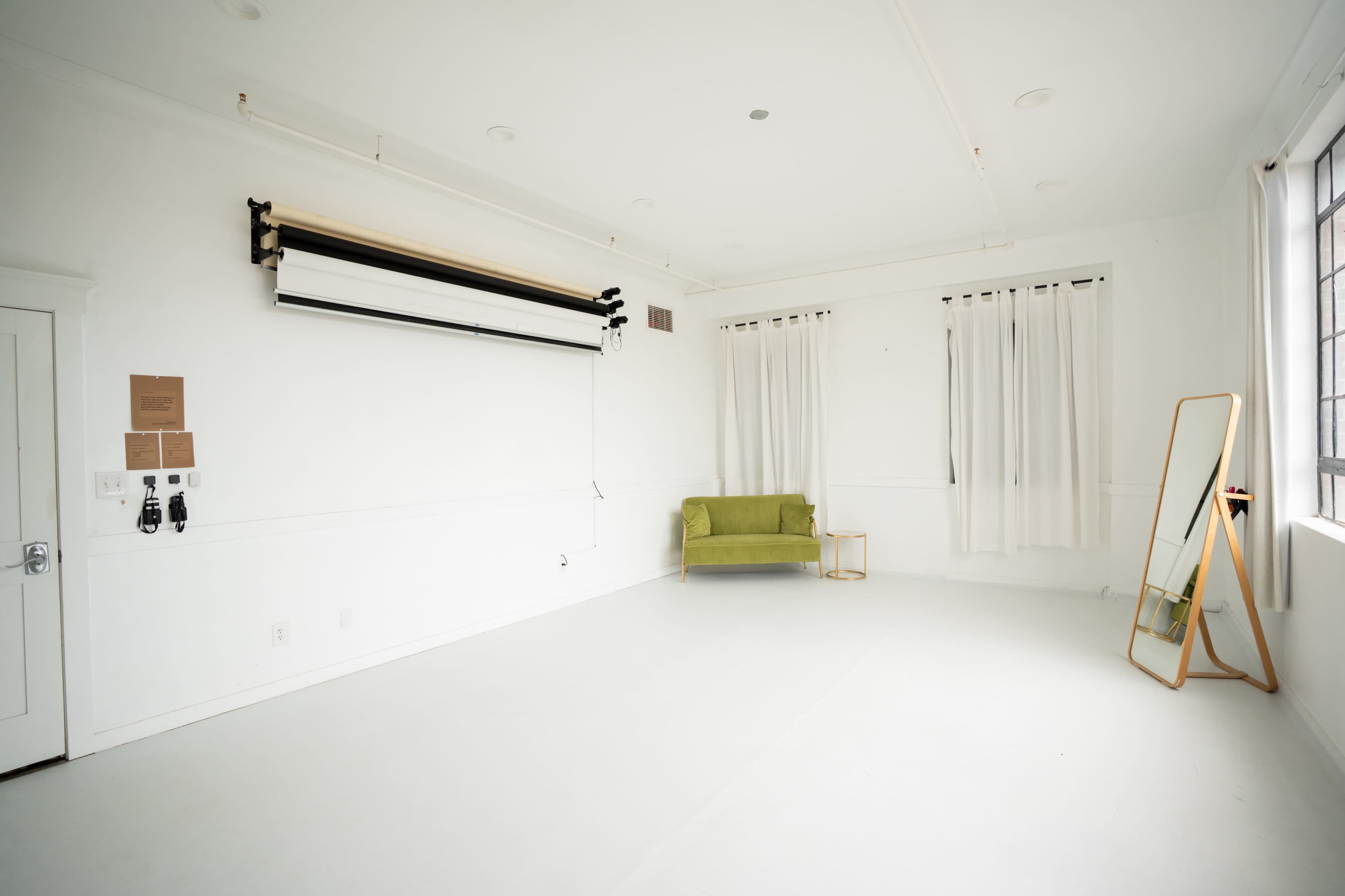 The image shows a bright, empty studio space featuring a green couch, a large mirror, and white walls with floor-to-ceiling curtains.