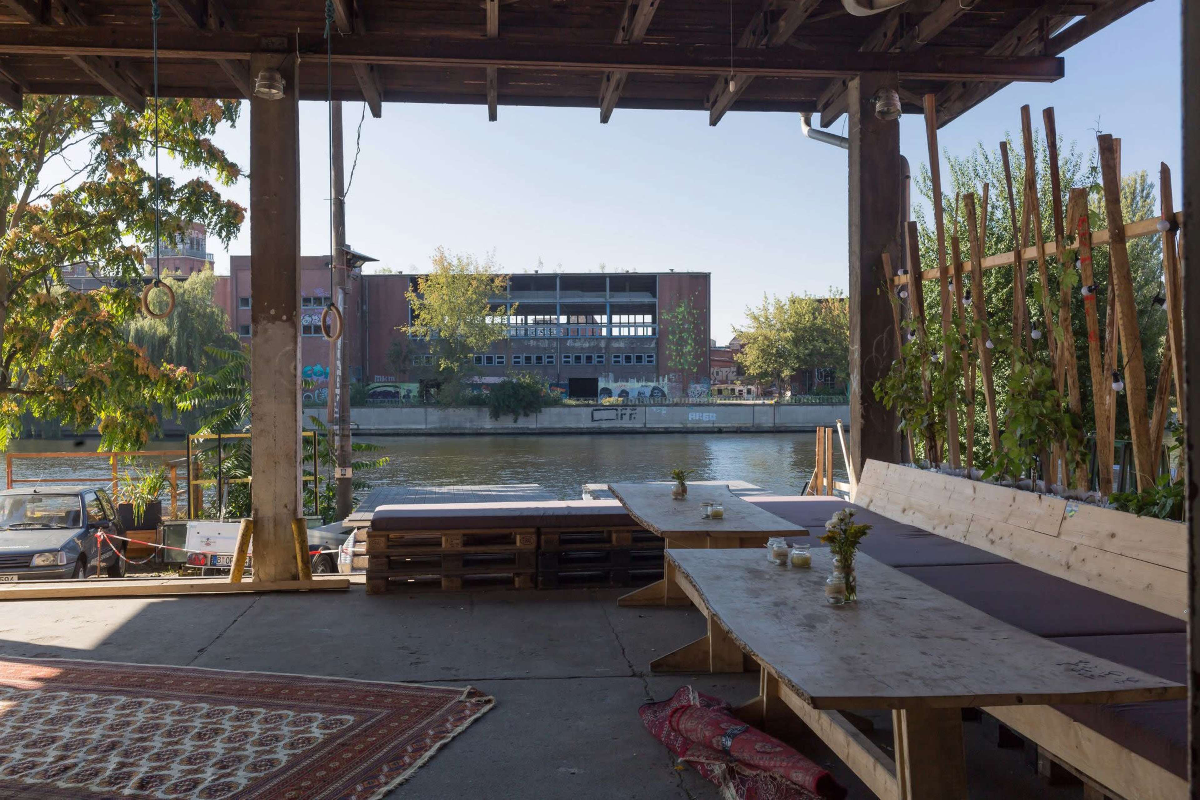 The scene shows an outdoor seating area made of wooden pallets next to a calm river, with a view of a brick building in the background.