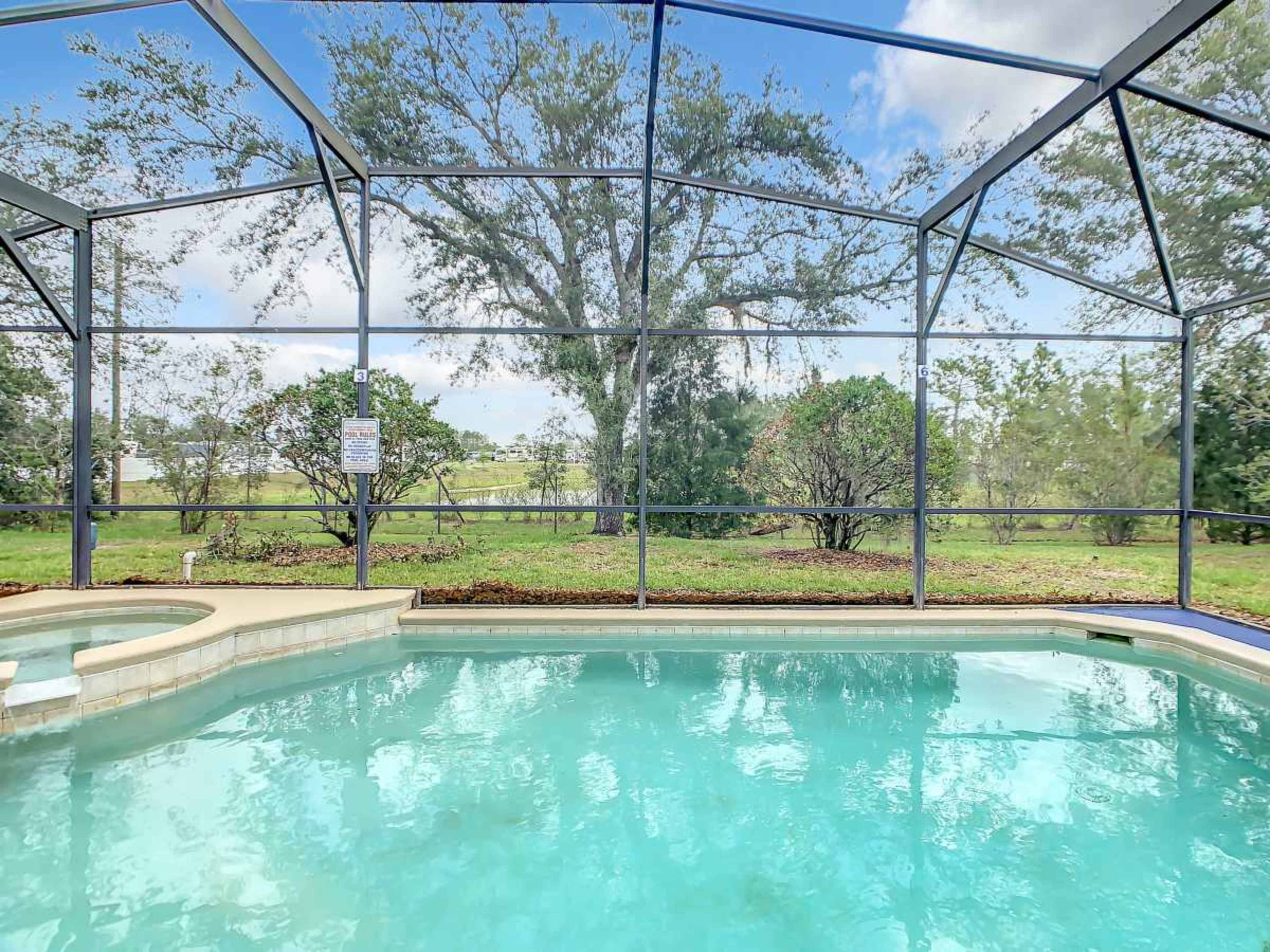 A clear swimming pool is enclosed by a screened patio, surrounded by green grass and trees in the background.