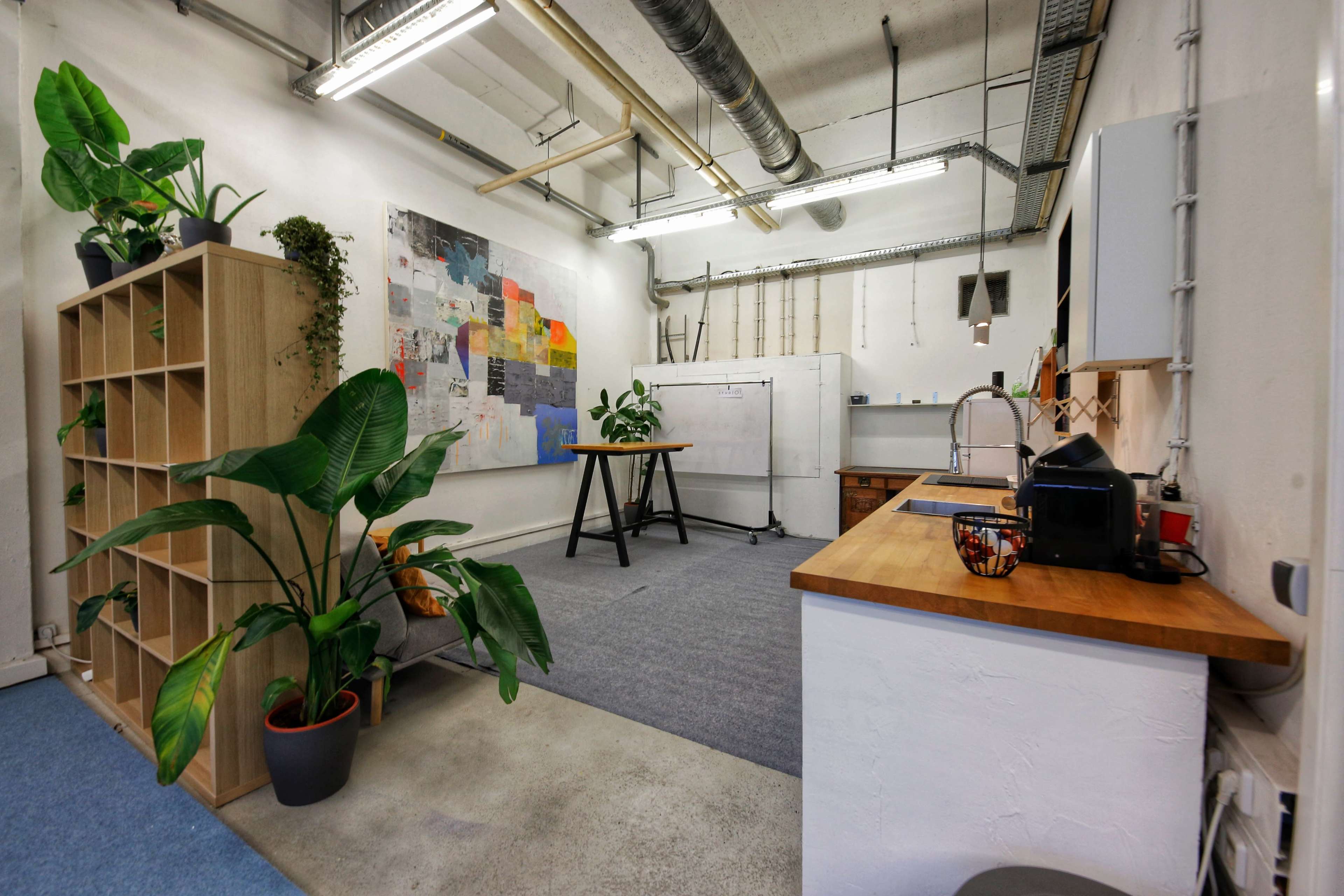 The image shows a minimalistic office space featuring a kitchenette, a wooden table, a large bulletin board, and several indoor plants.