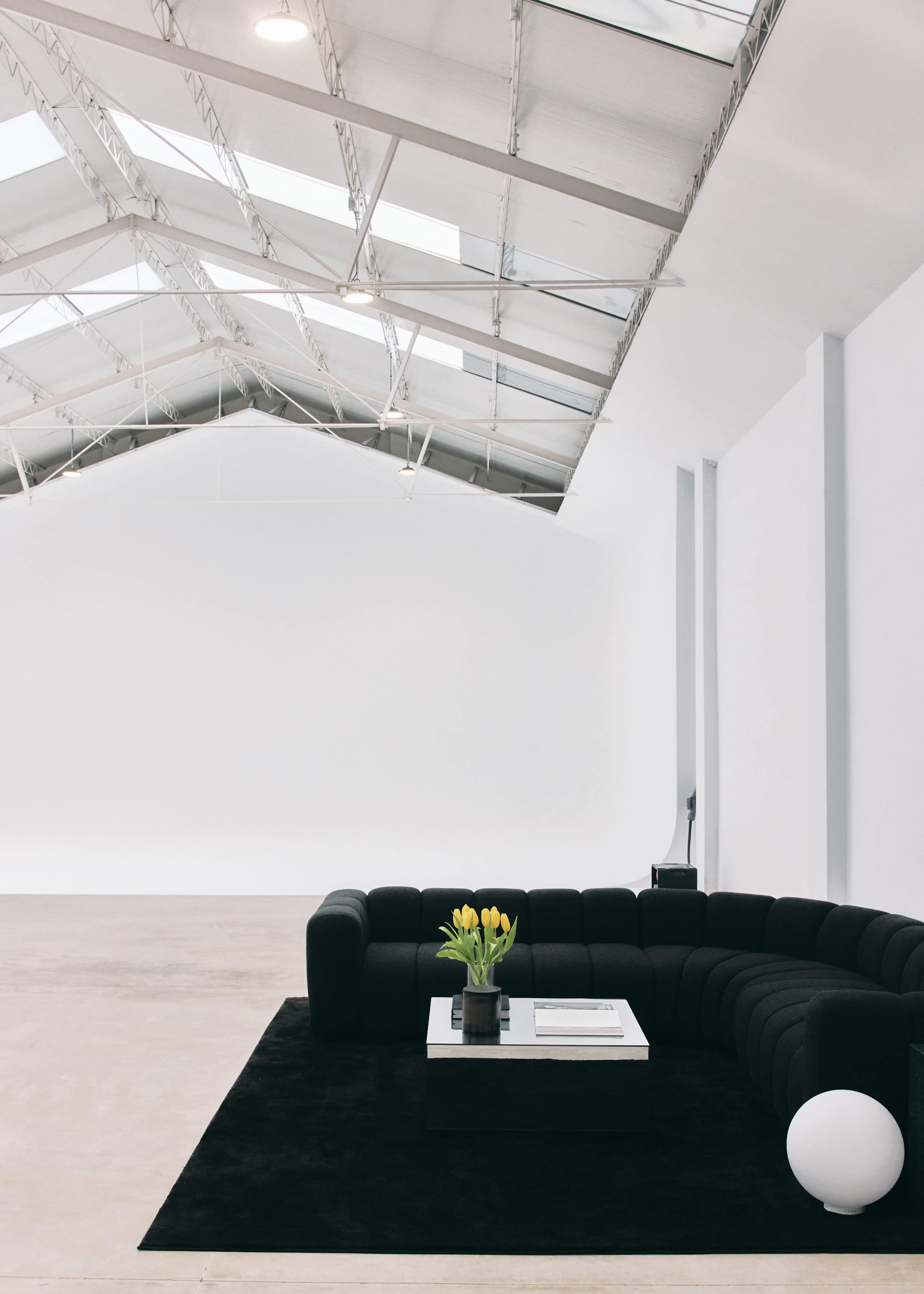 A modern interior space features a black sectional sofa, a glass coffee table with a vase of yellow tulips, and a large white spherical lamp on a black rug.