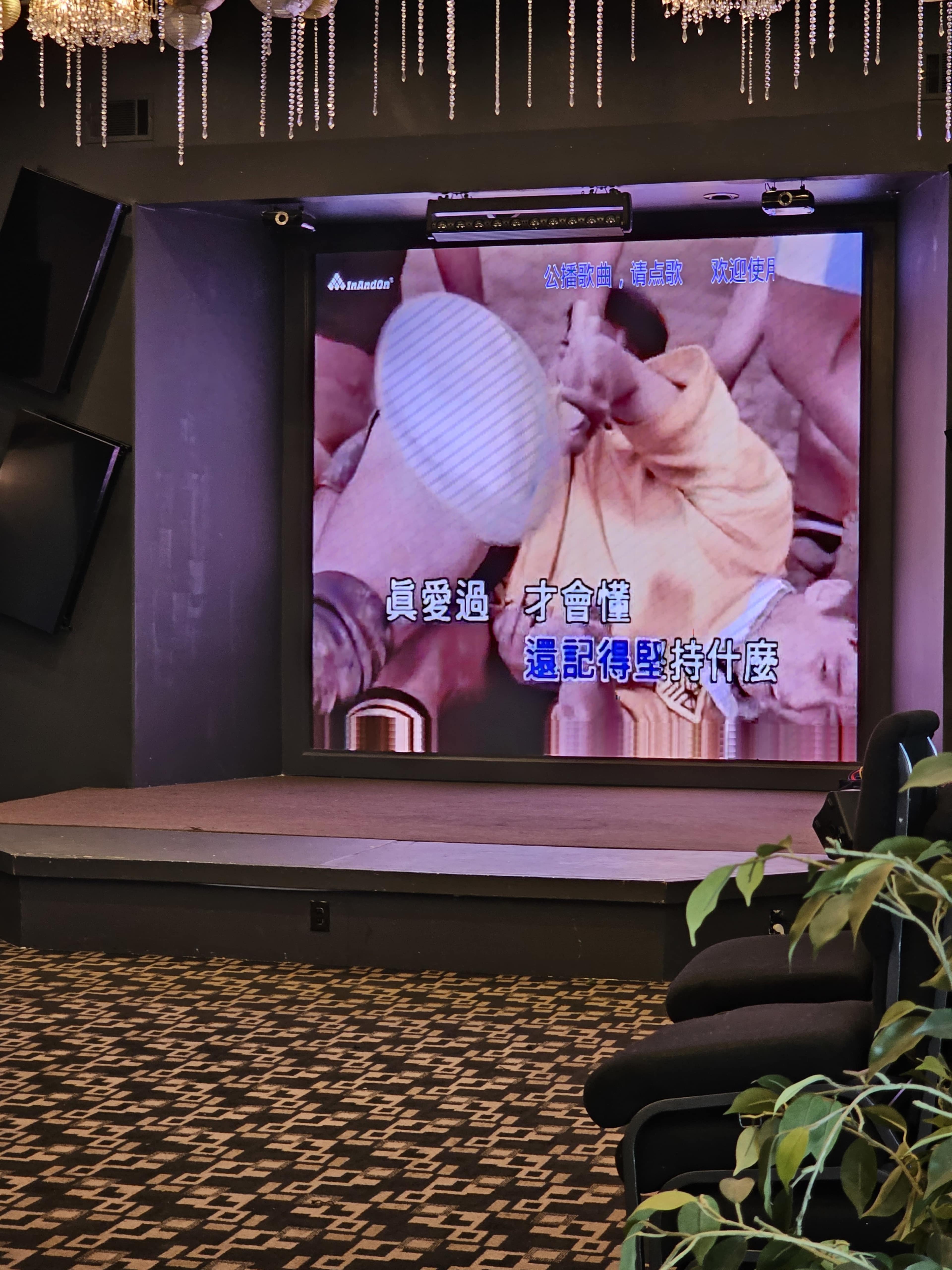 A large screen displaying karaoke lyrics while a cozy seating area is visible in the foreground.