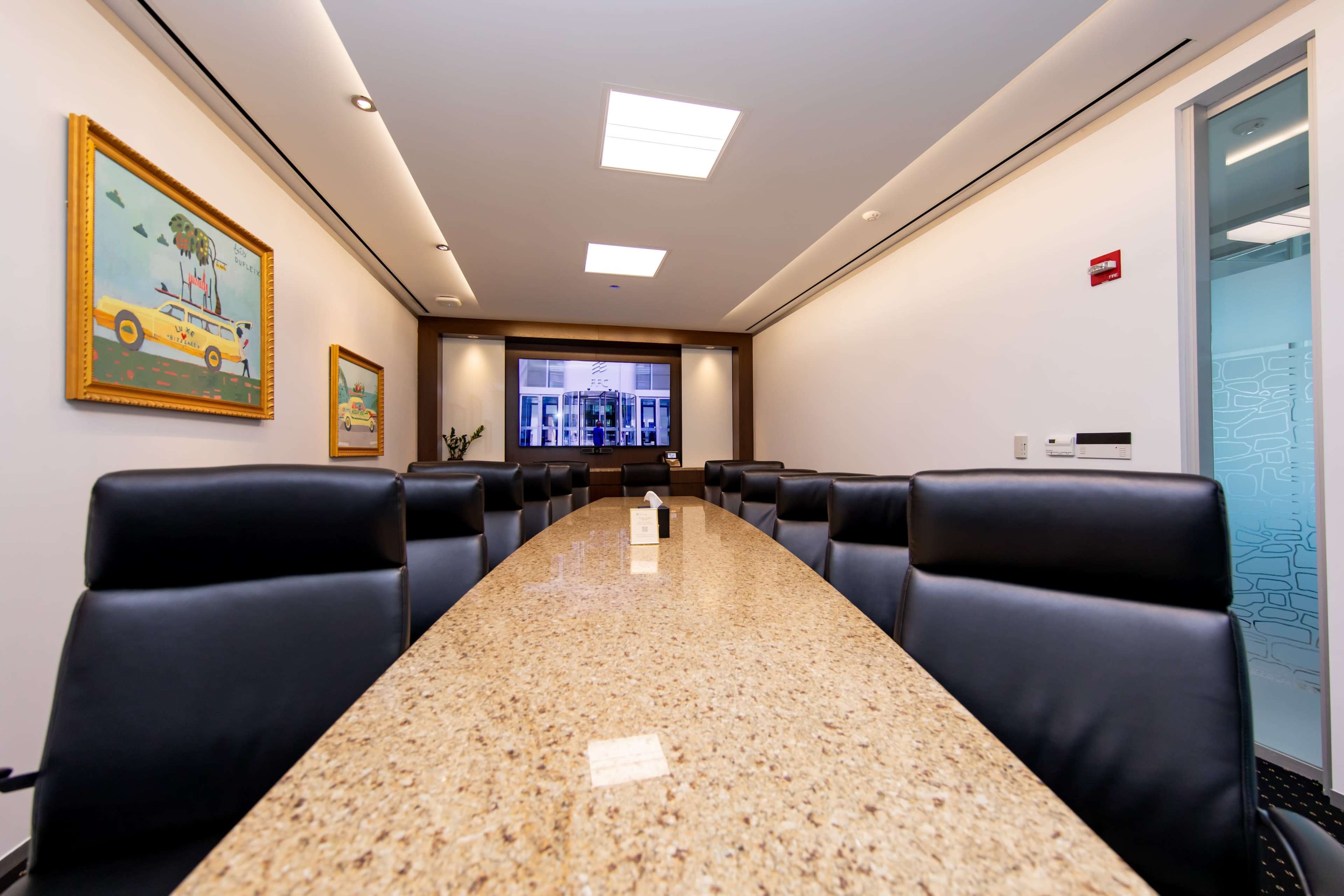 A long, oval conference table with black leather chairs is set in a bright, modern meeting room featuring a large window and a piece of artwork on the wall.