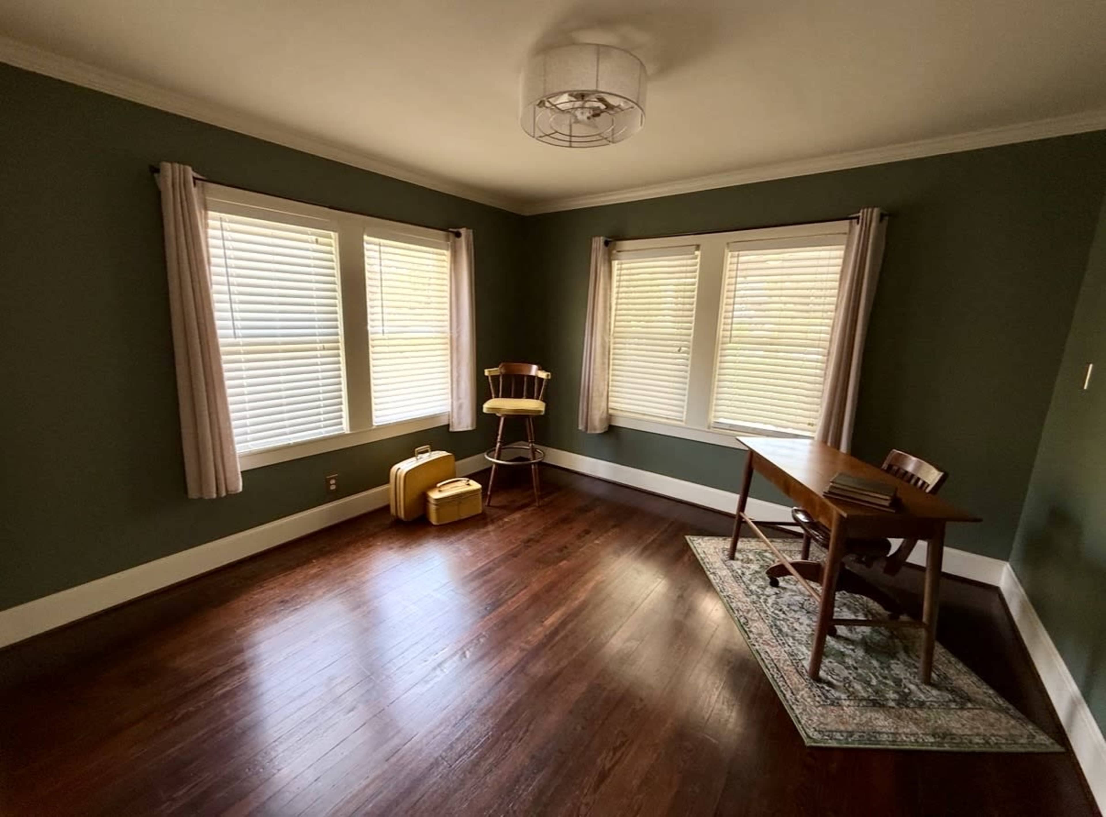 The image depicts a small, empty room with green walls, a wooden table, a chair, and two windows adorned with light-colored curtains.