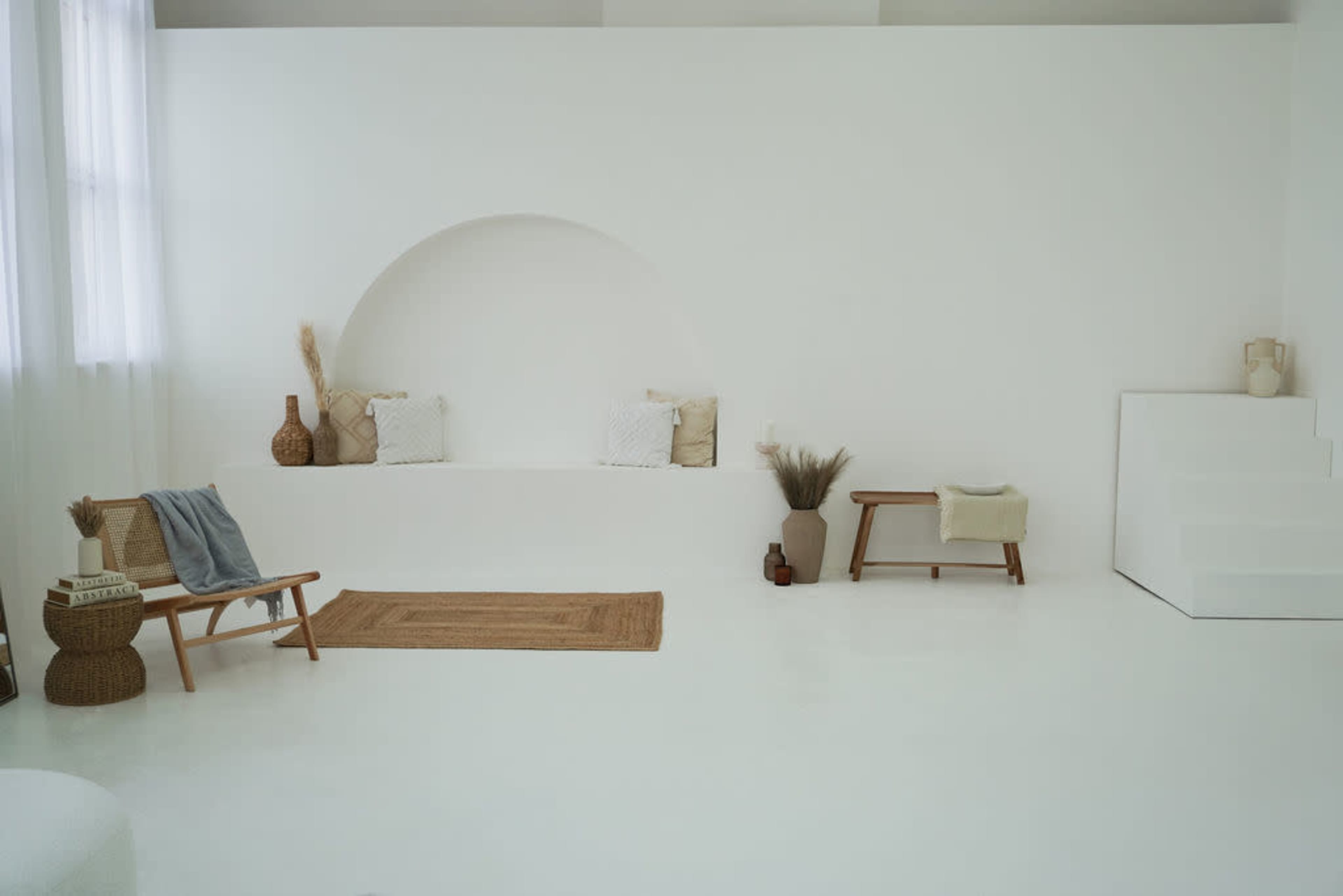 The image shows a minimalist interior with white walls, a curved alcove with cushions, a woven rug on the floor, and simple furniture arranged in the space.