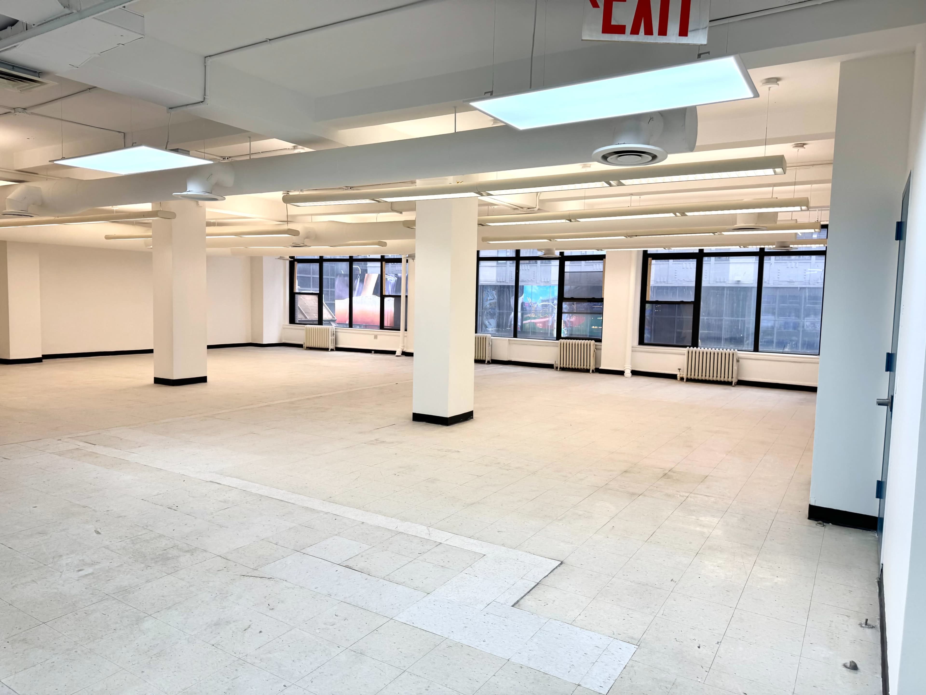 The image shows an empty office space with large windows, fluorescent lighting, and a light-colored tiled floor.
