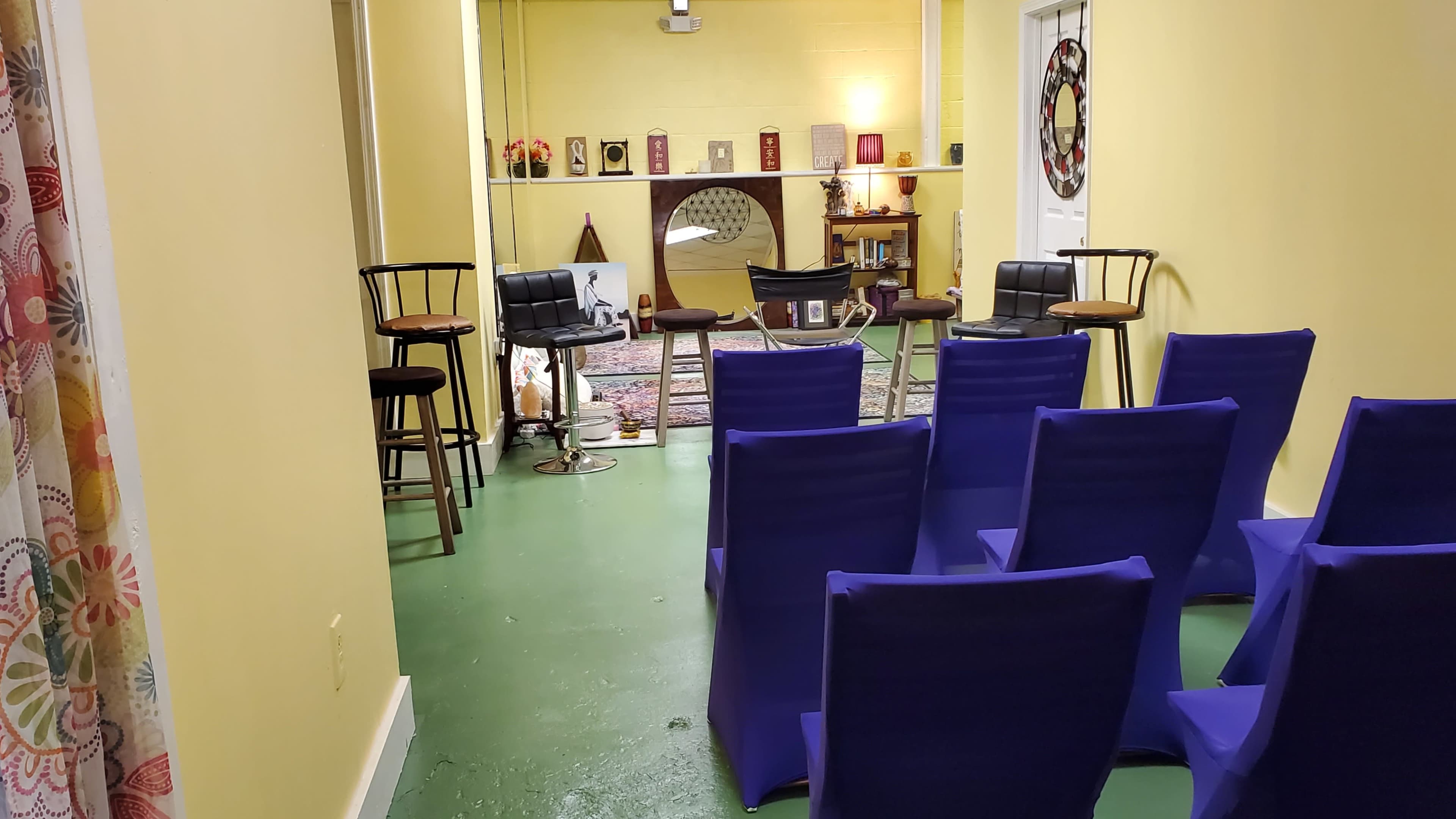 The image shows a small room with yellow walls, arranged with chairs and bar stools facing a central area that features a decorative mirror and various framed items.