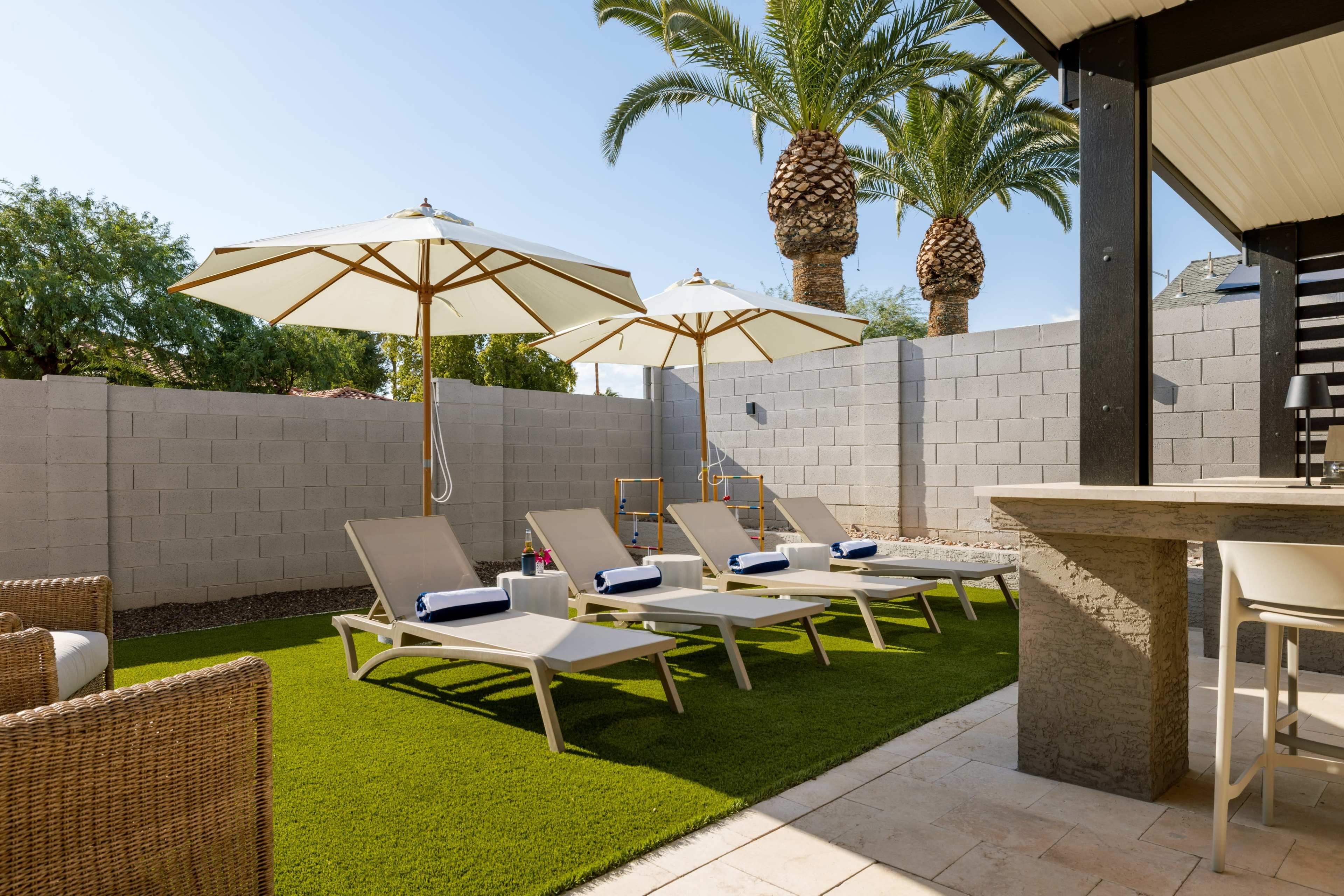 The image shows a backyard with several lounge chairs and umbrellas arranged on green artificial grass, surrounded by palm trees and a gray block wall.