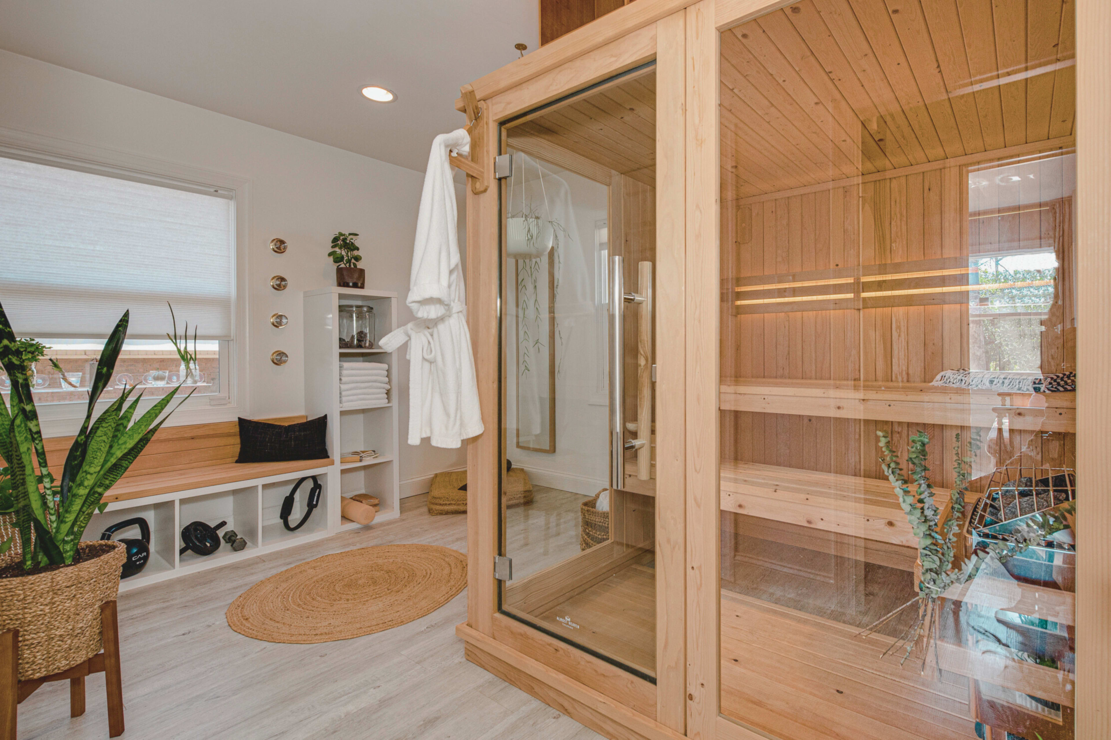 A modern bathroom featuring a wooden sauna, a plant, a white robe hanging on the door, and organized shelves with towels.