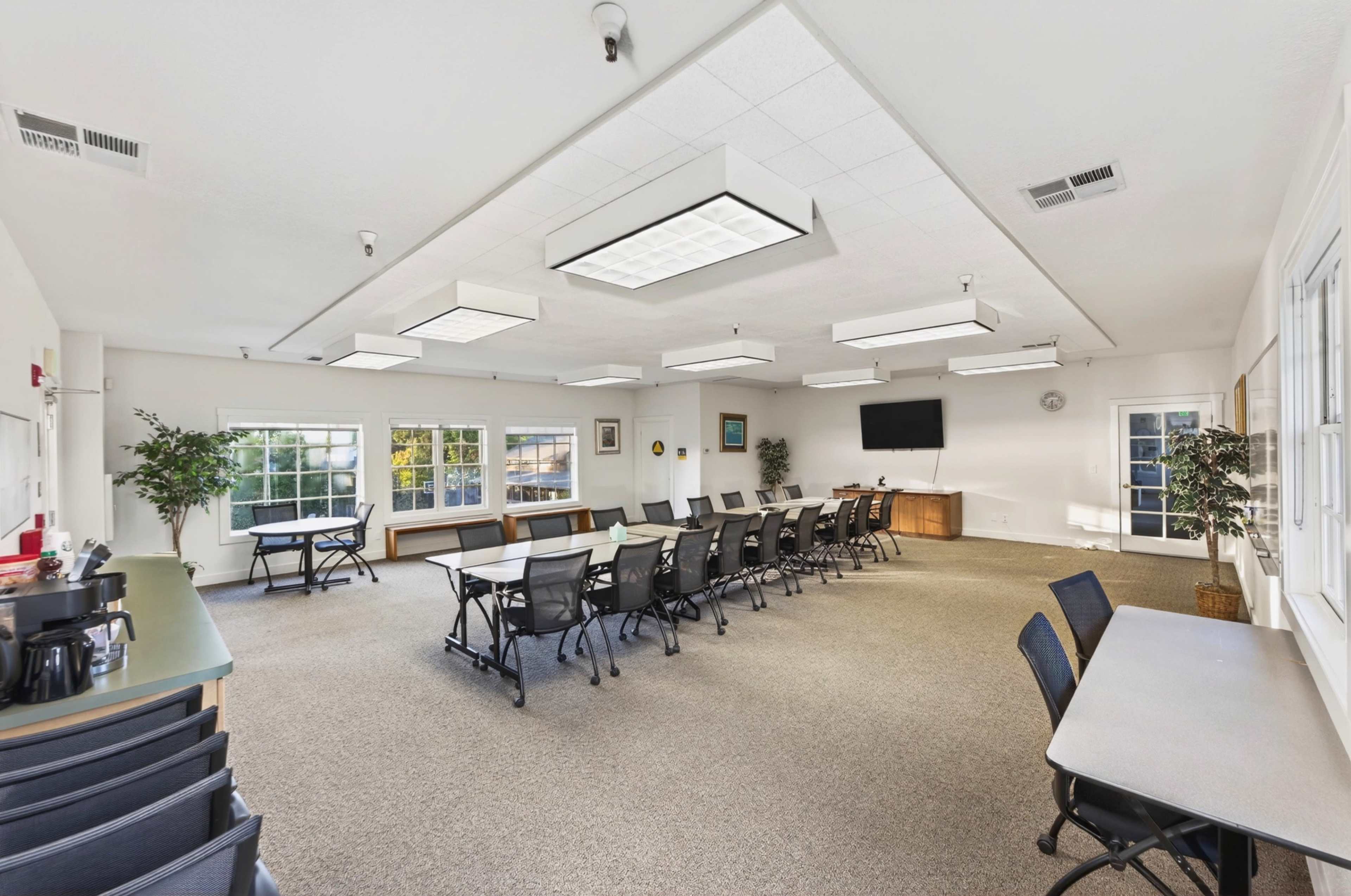 The image shows a spacious meeting room with tables arranged in a U-shape, surrounded by large windows and equipped with overhead lighting and a presentation screen.
