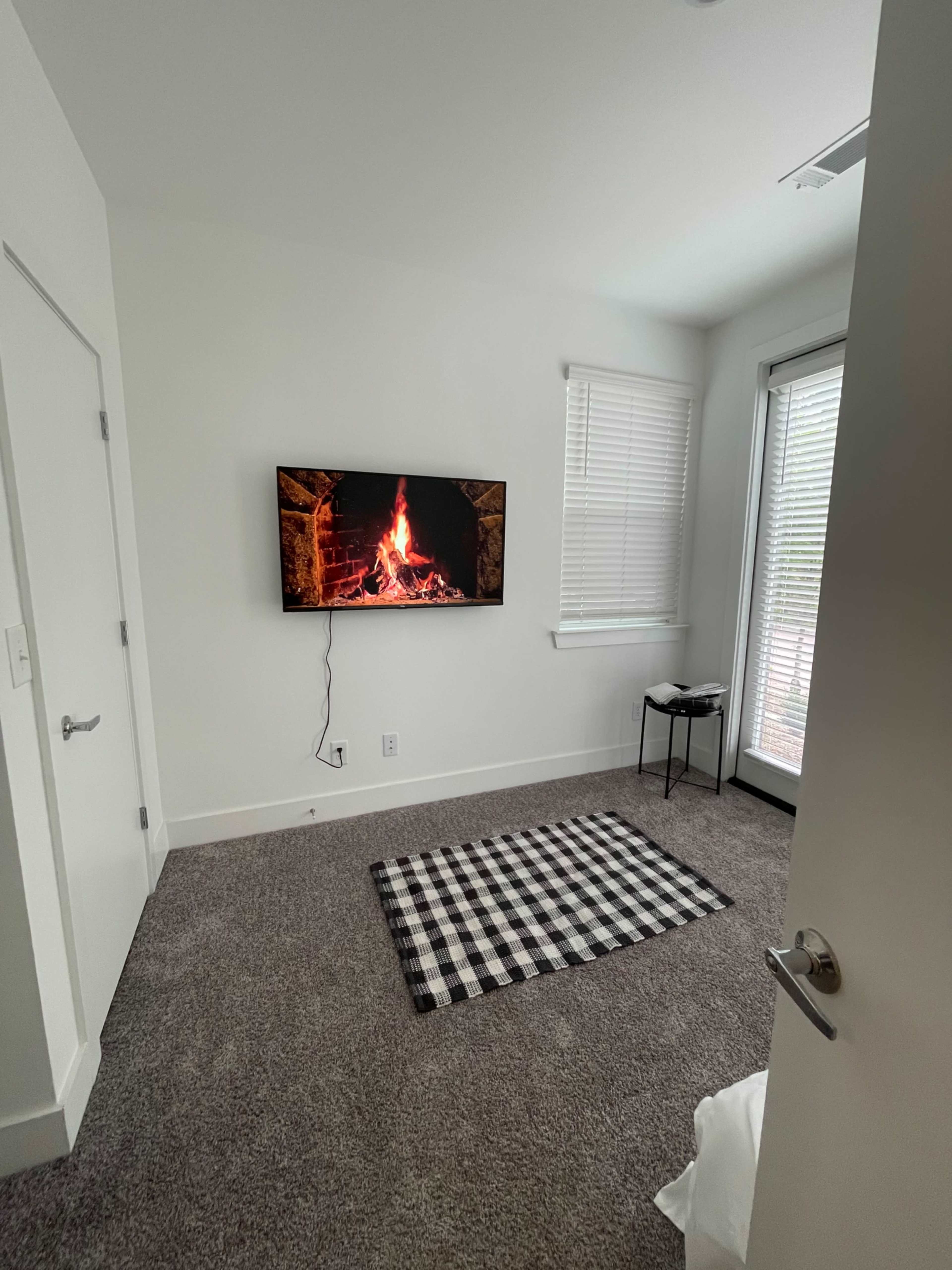 A cozy room features a flat-screen TV displaying a fireplace, a checkered rug, and a door opening to a balcony.