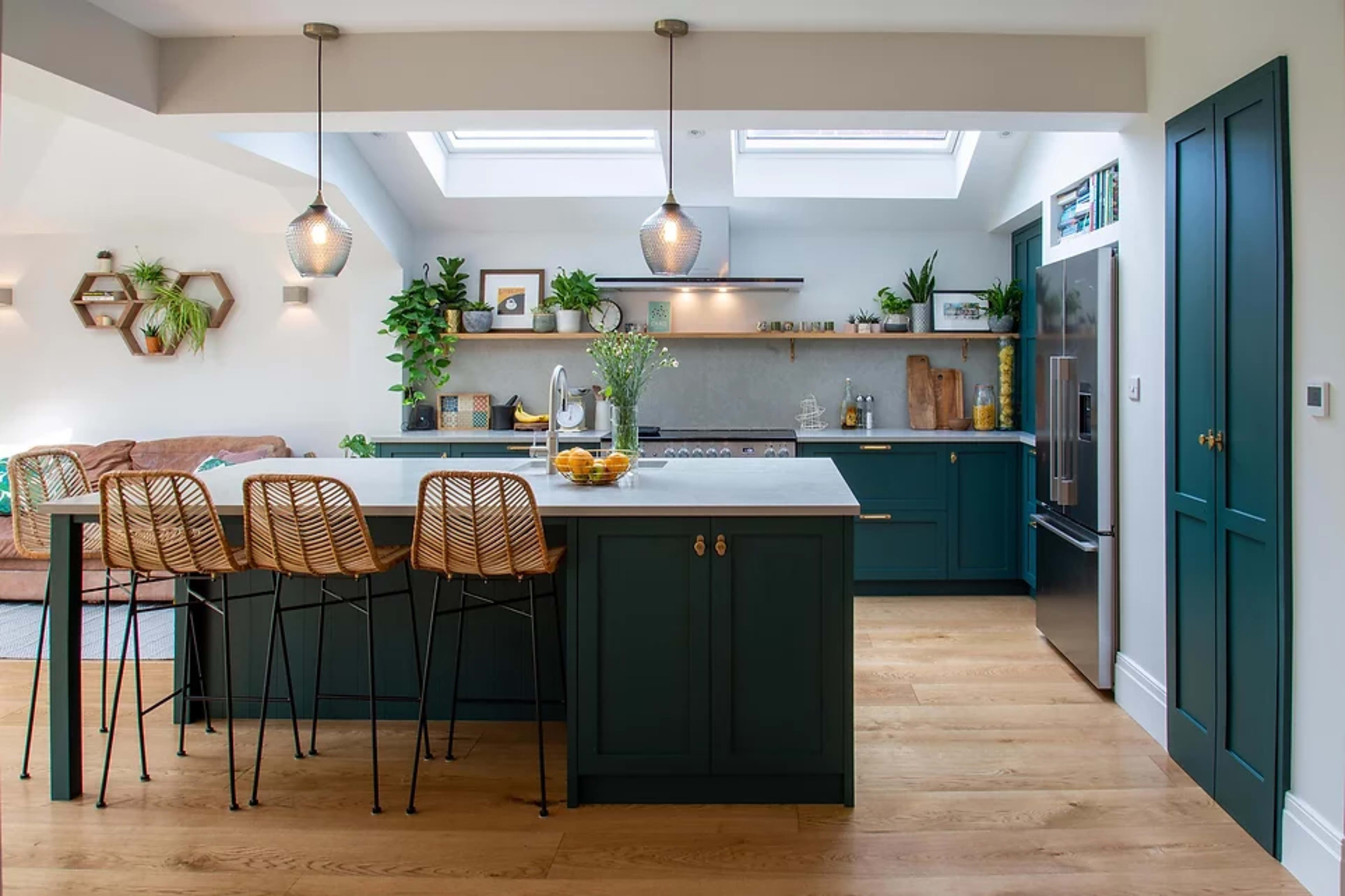 A modern kitchen with a central island, bar stools, and skylights, featuring green cabinetry and plants.