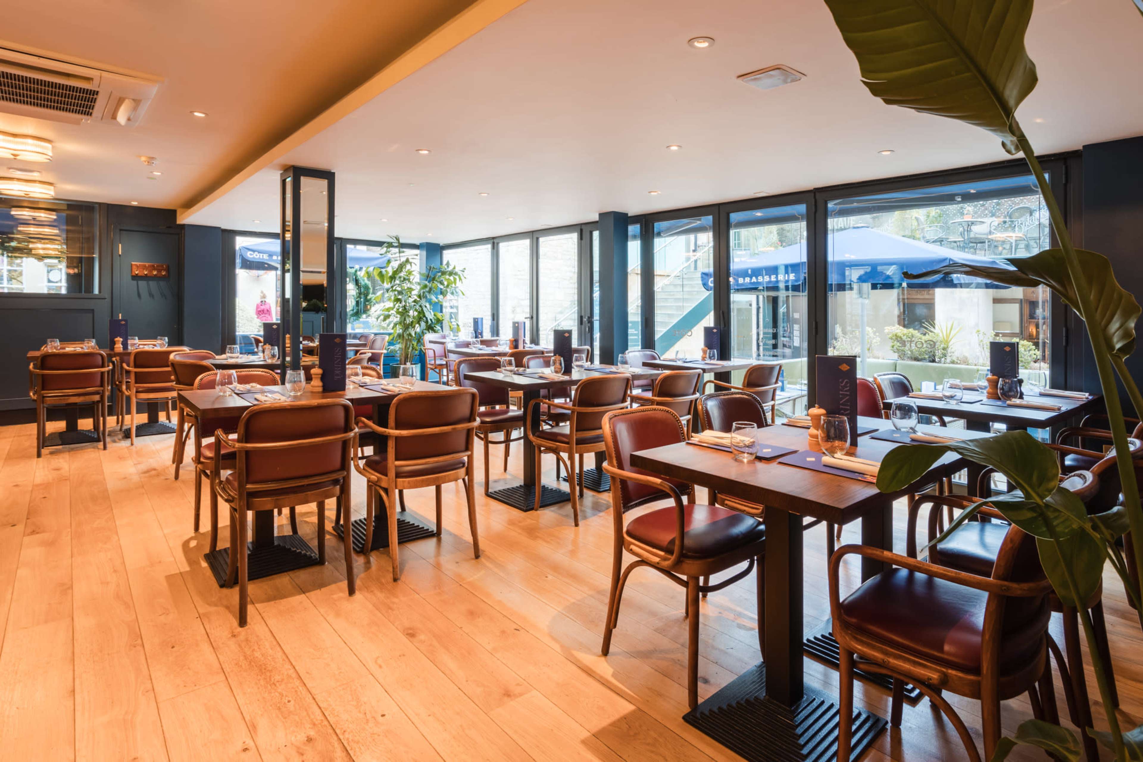 The image shows a modern restaurant interior with wooden floors, neatly arranged tables, and large windows allowing natural light to enter.