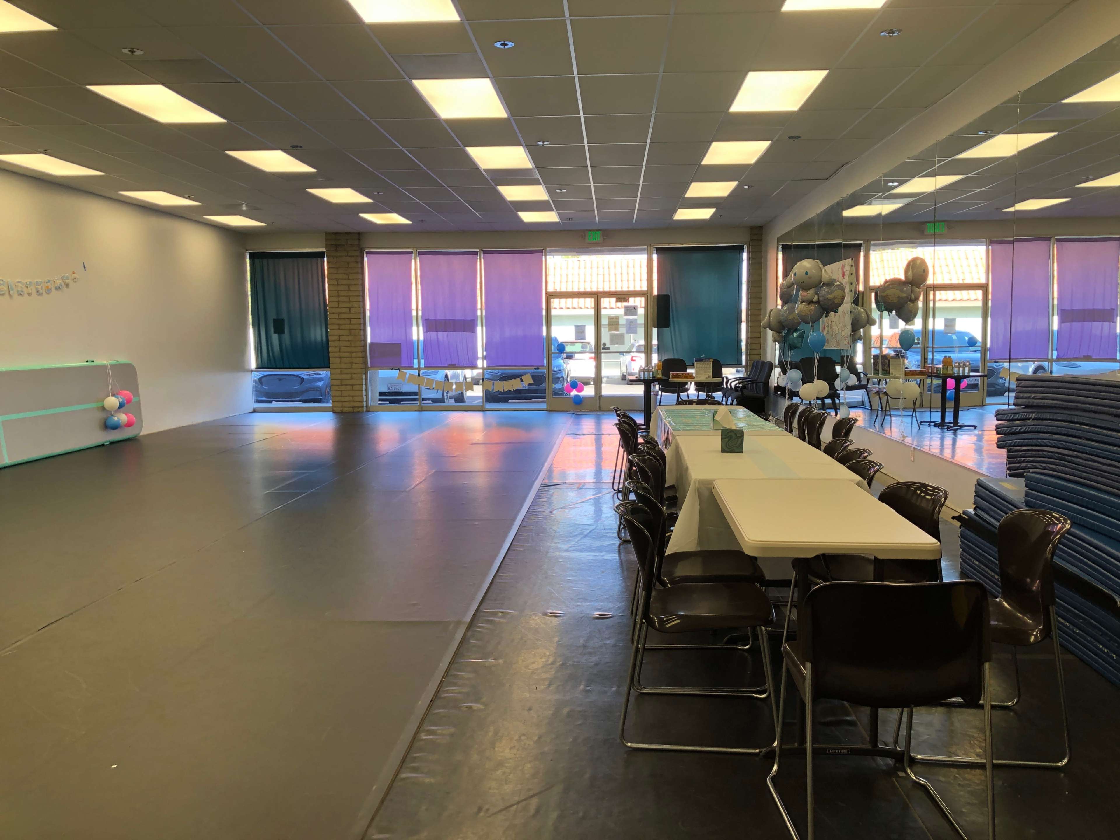 The image shows a spacious event room with a long table and chairs arranged in the center, surrounded by mirrors and decorated with balloons near the entrance.
