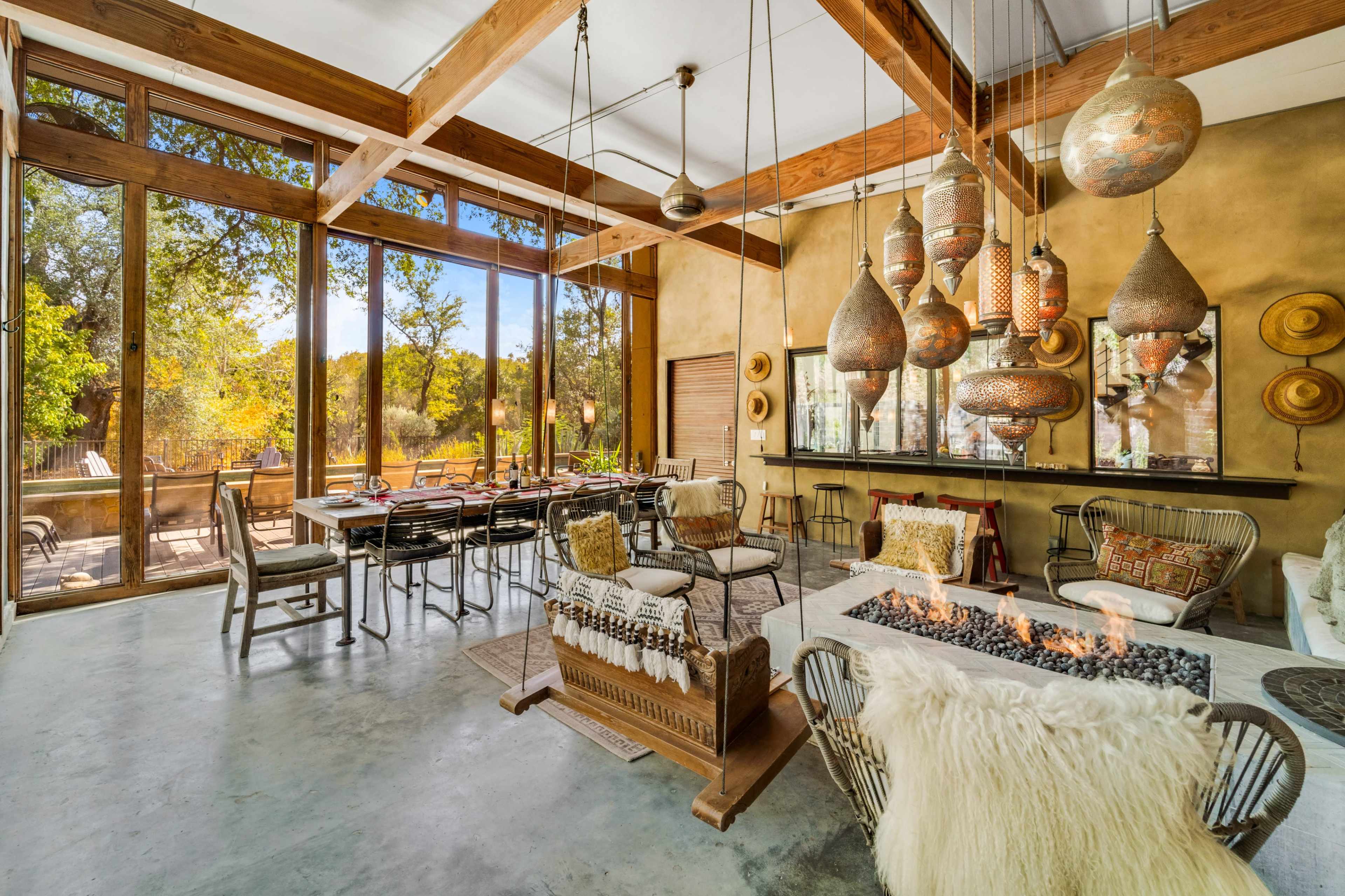 The image shows a modern interior with large windows allowing natural light, featuring a dining area, comfortable seating, and decorative hanging lamps.