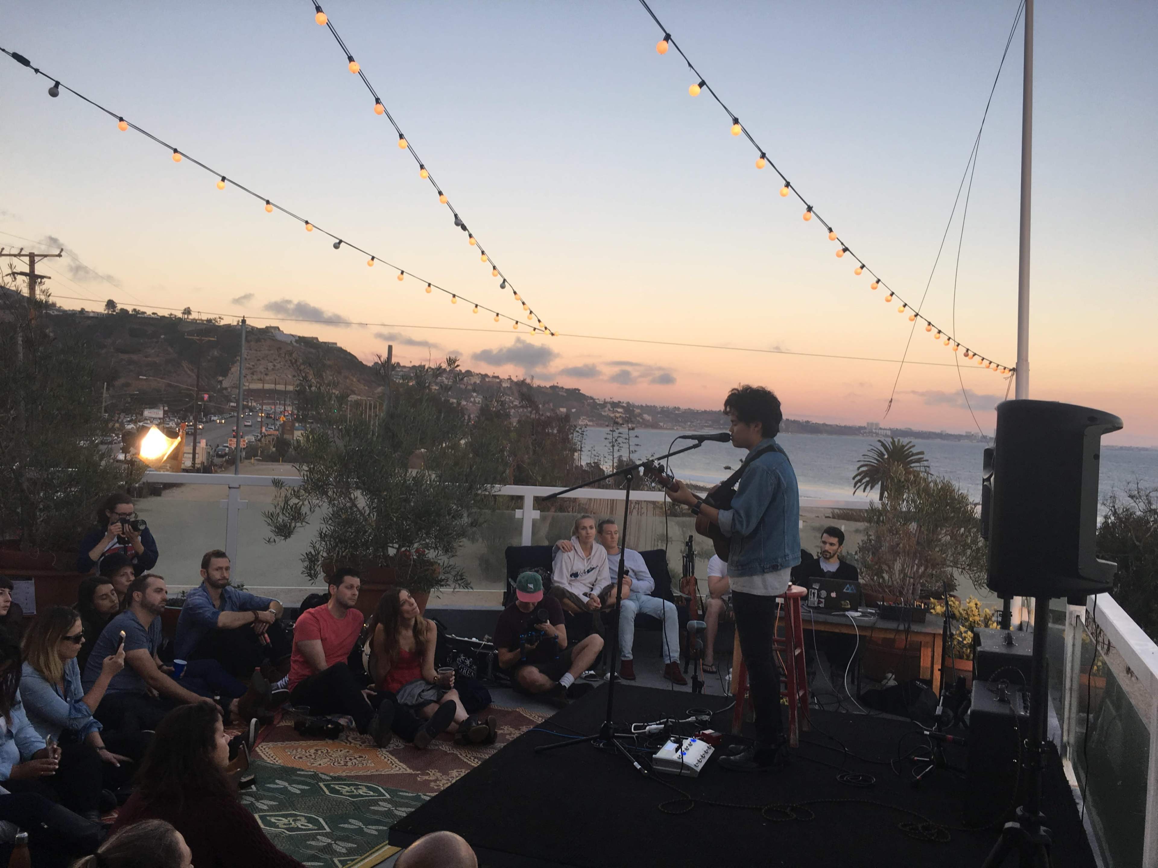 A musician performs on a rooftop stage as an audience sits on the ground, overlooking the ocean at sunset.