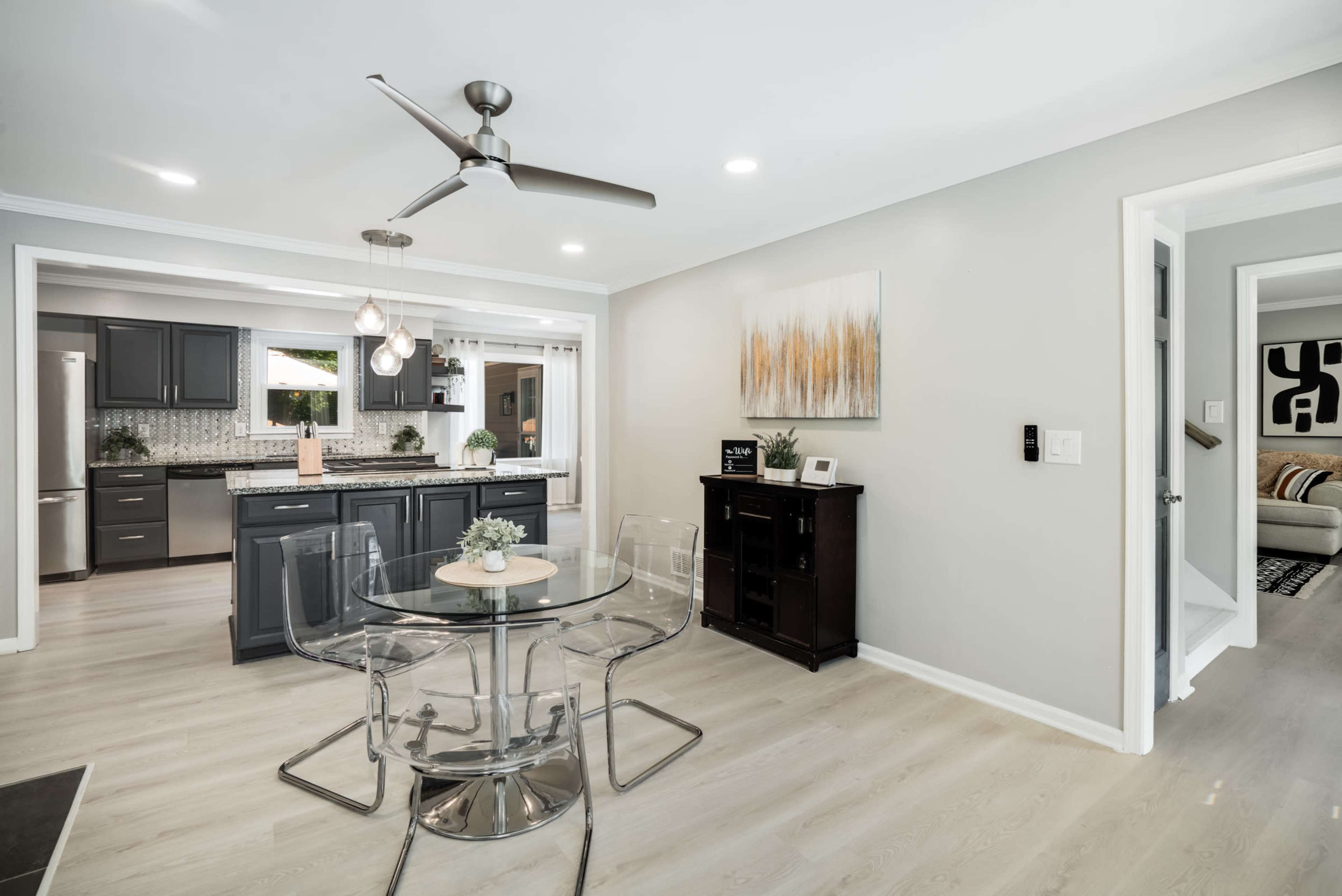 A modern dining area with a round glass table and clear chairs, adjacent to a kitchen featuring dark cabinetry and stainless steel appliances.
