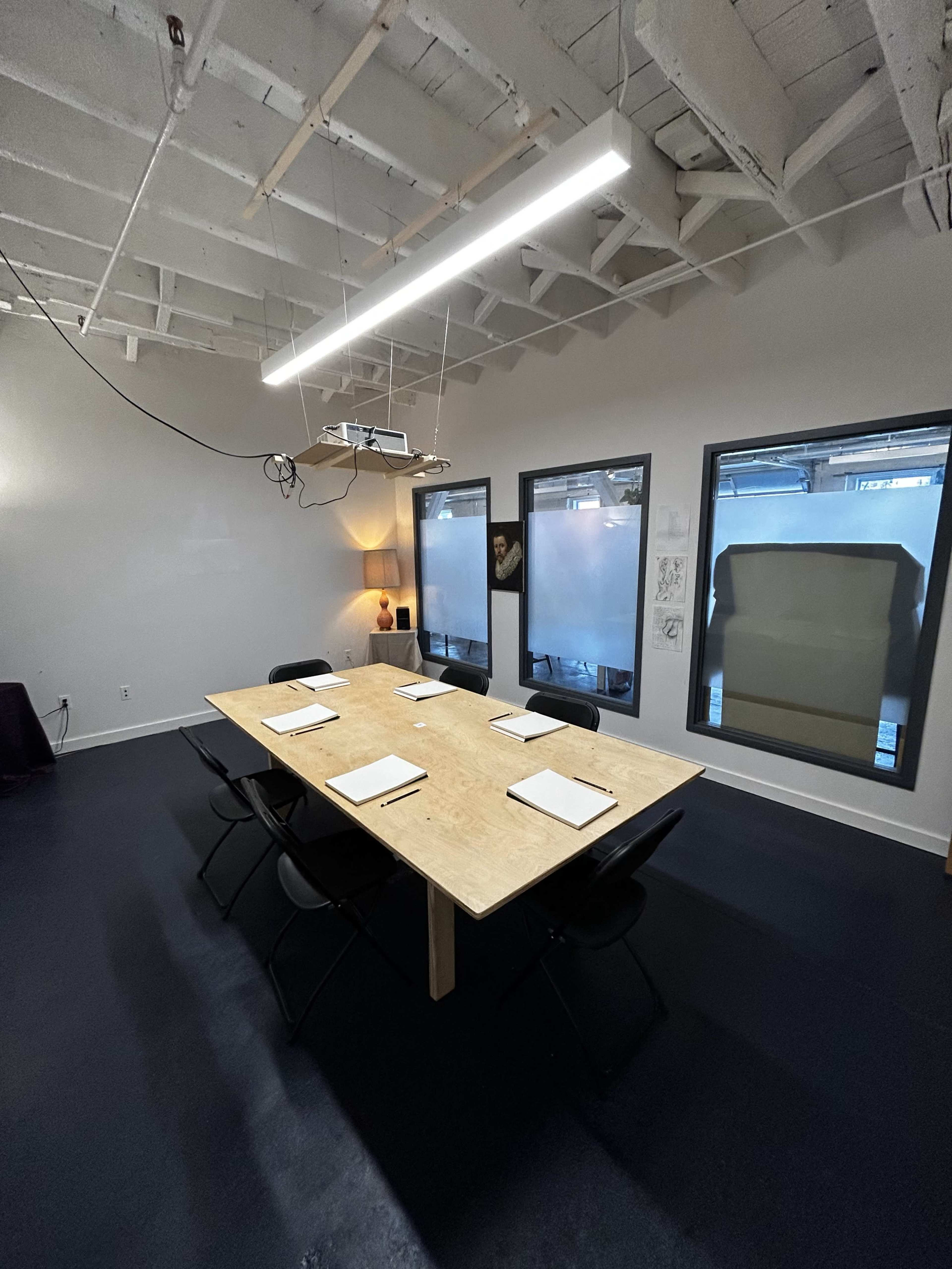 A long wooden table with chairs is arranged in a modern conference room featuring a ceiling with exposed beams and large windows.