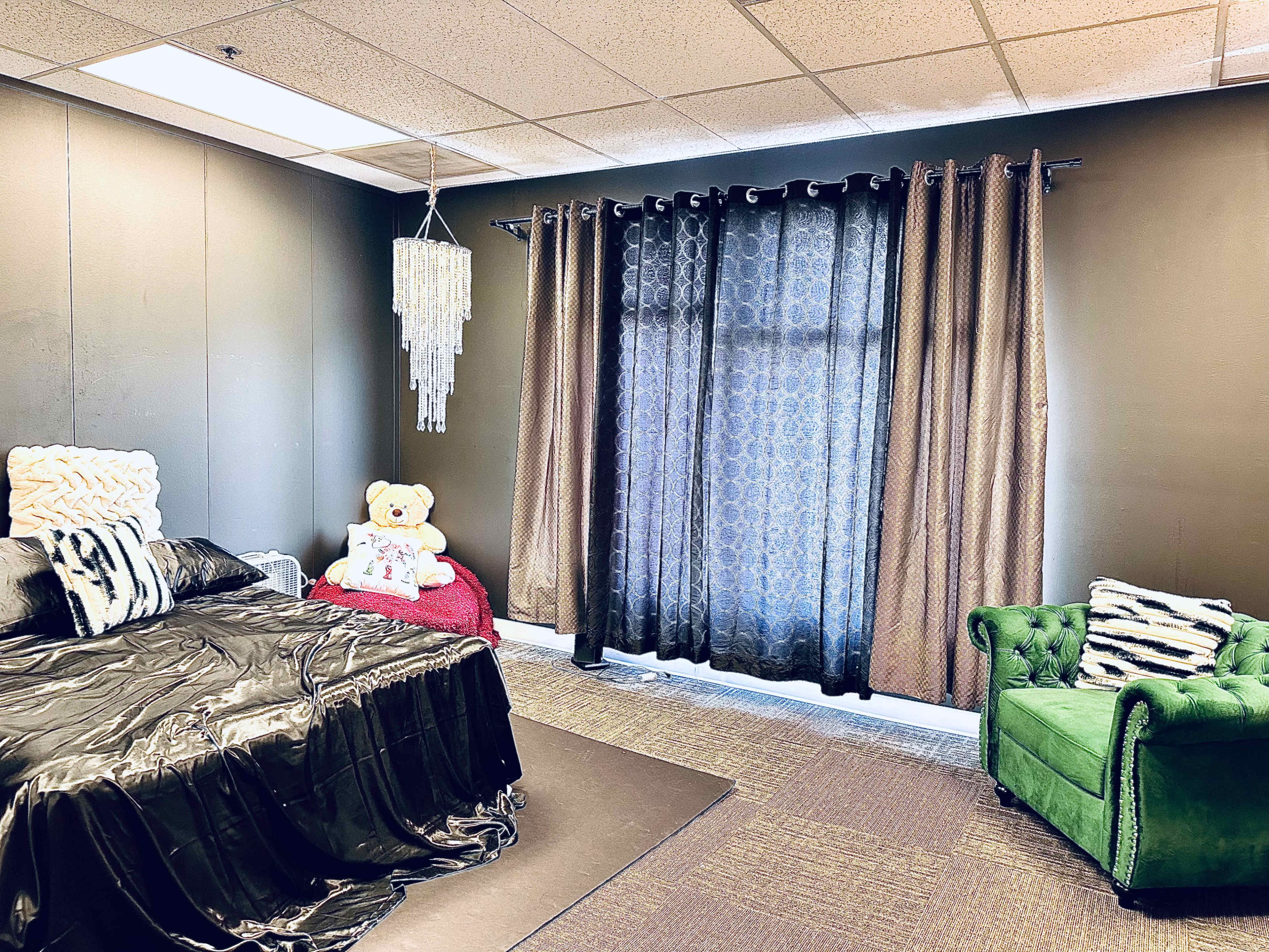 A bedroom featuring a black bedspread, a teddy bear on the bed, a green chair, and dark curtains covering a window.