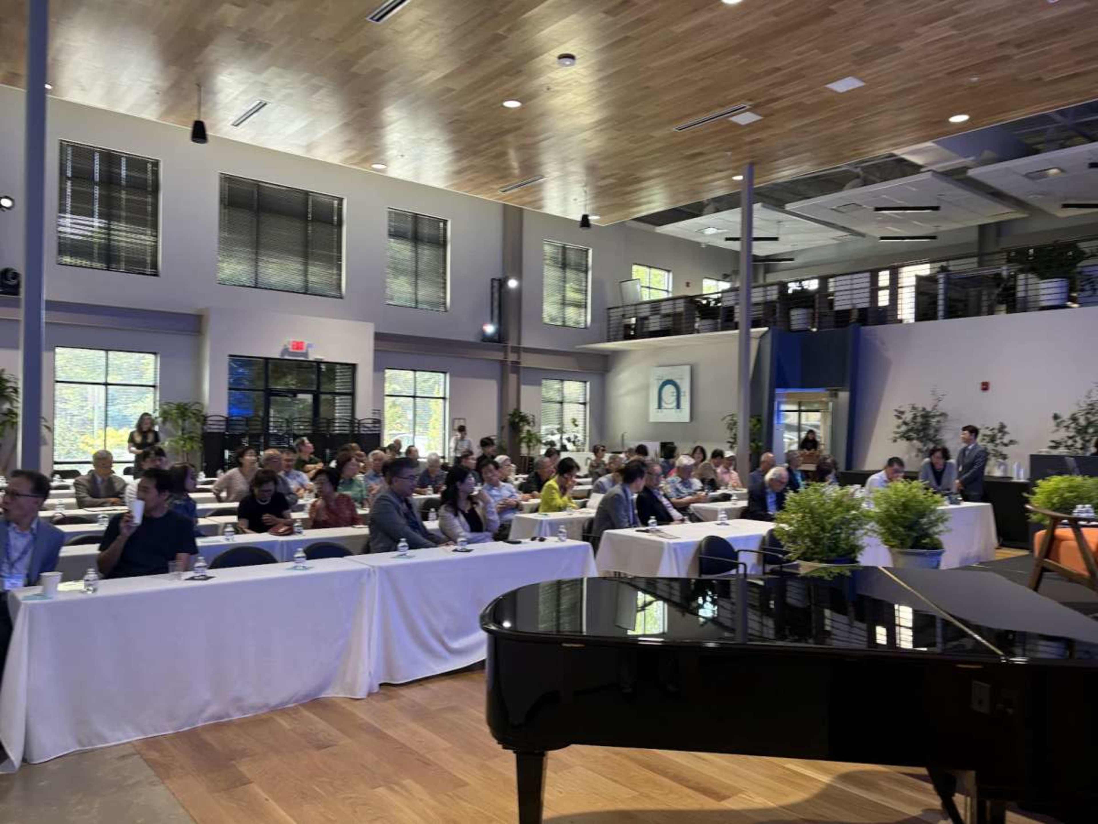 A group of attendees sits in a spacious conference room with large windows, rows of tables, and a grand piano in the foreground.