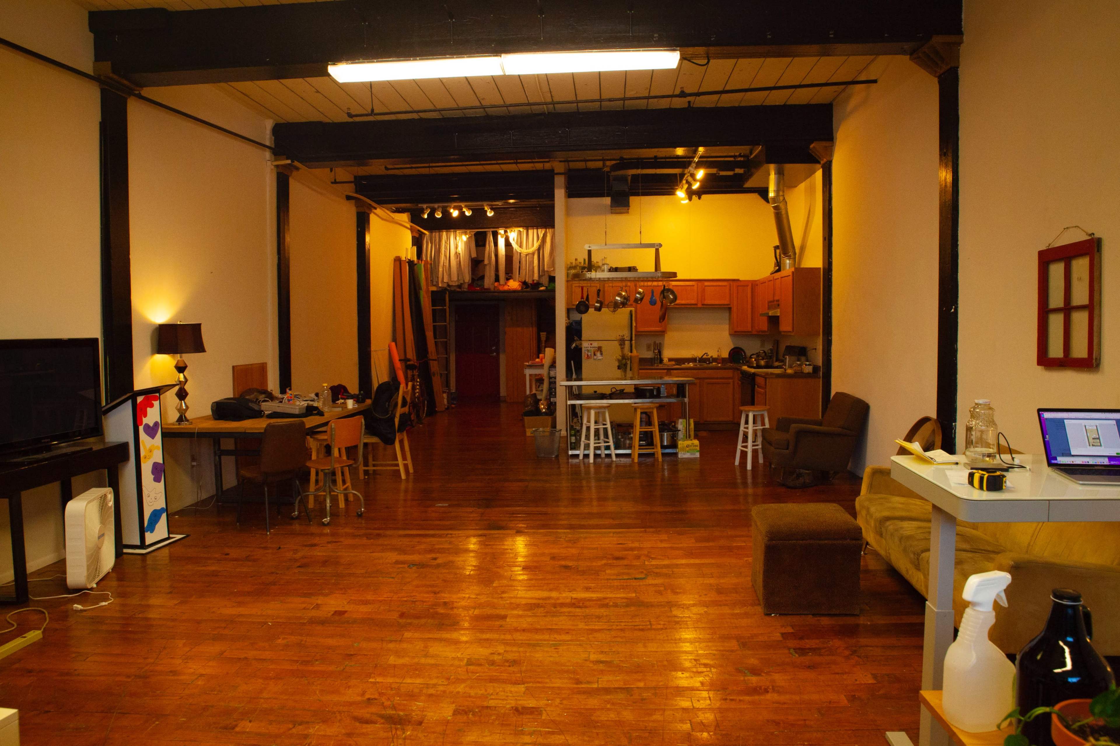 The image shows a spacious, open-plan interior with wooden flooring, featuring a kitchen area, dining space with tables and chairs, and a living area with a couch and television.