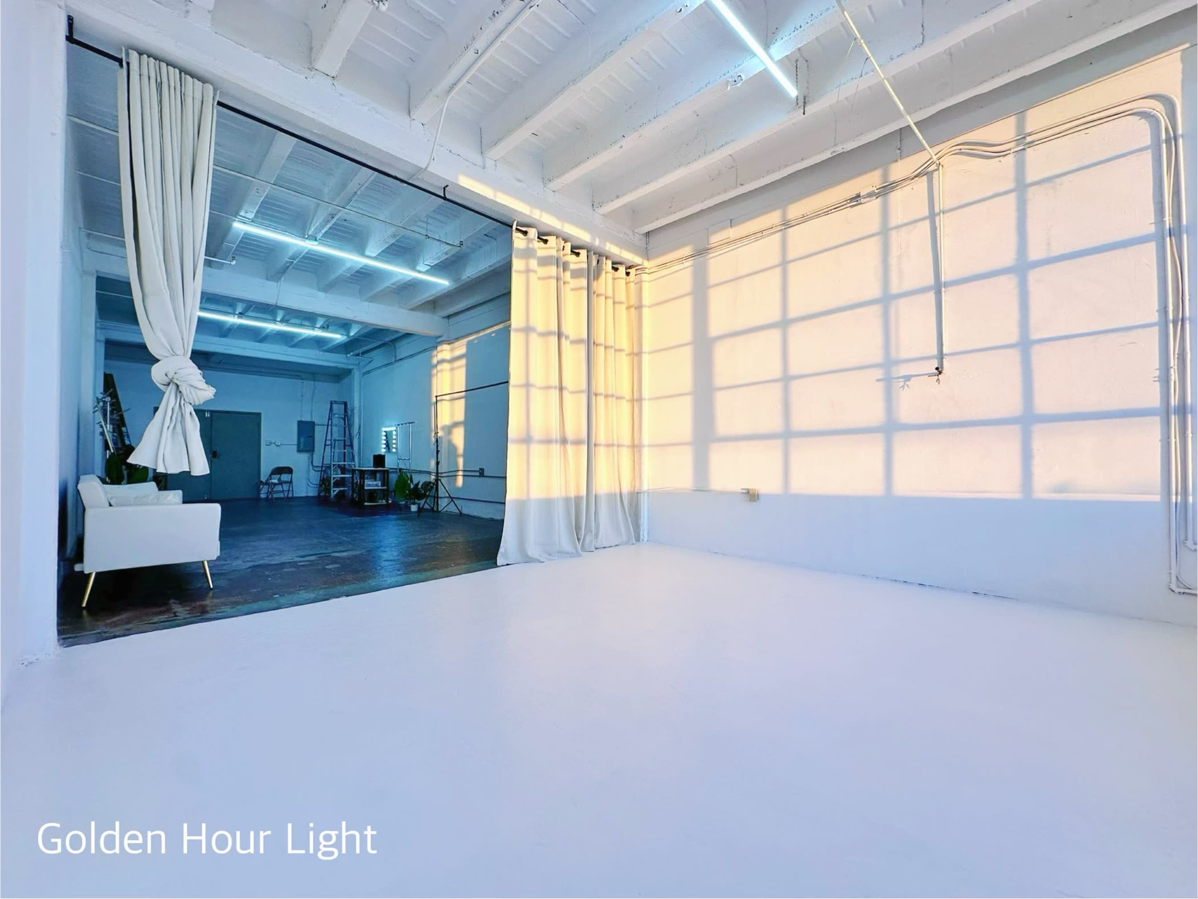 The image shows a spacious, well-lit room with sunlight casting long shadows across white walls and a minimalistic decor featuring a curtain and a small seating area.