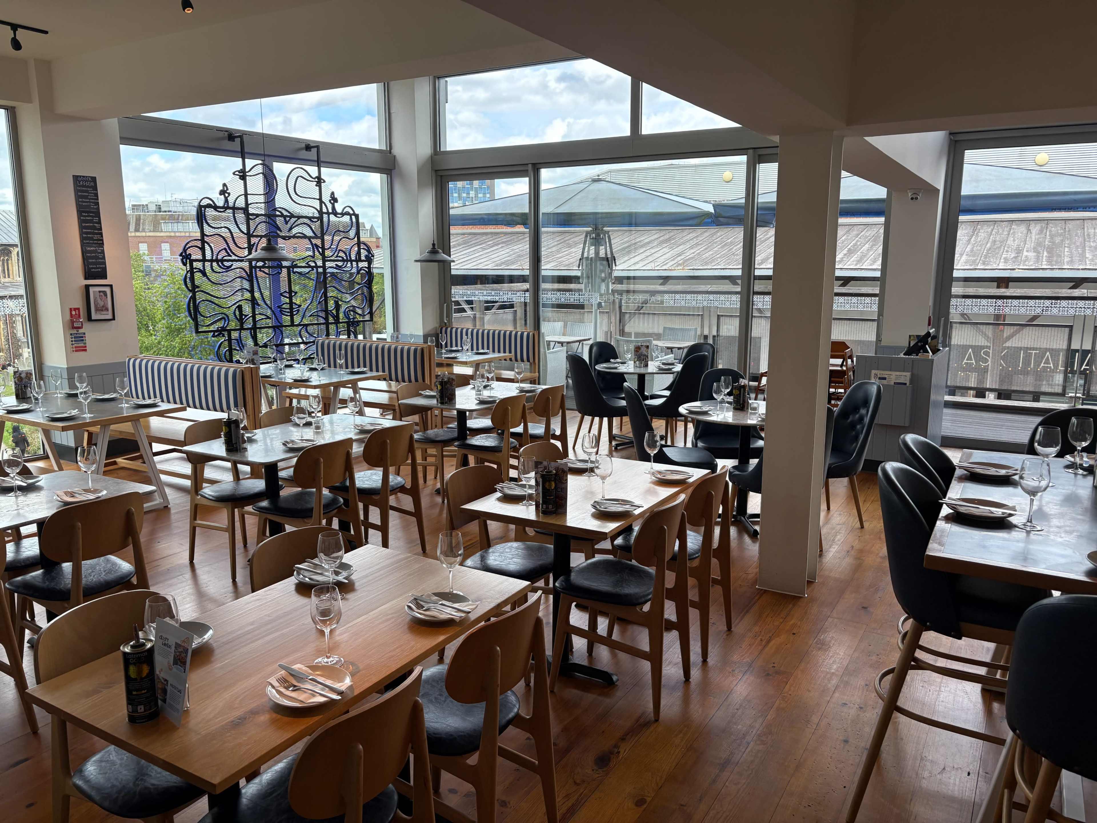 The image shows a brightly lit restaurant interior with wooden floors, tables set for dining, and large windows overlooking a view of a building.