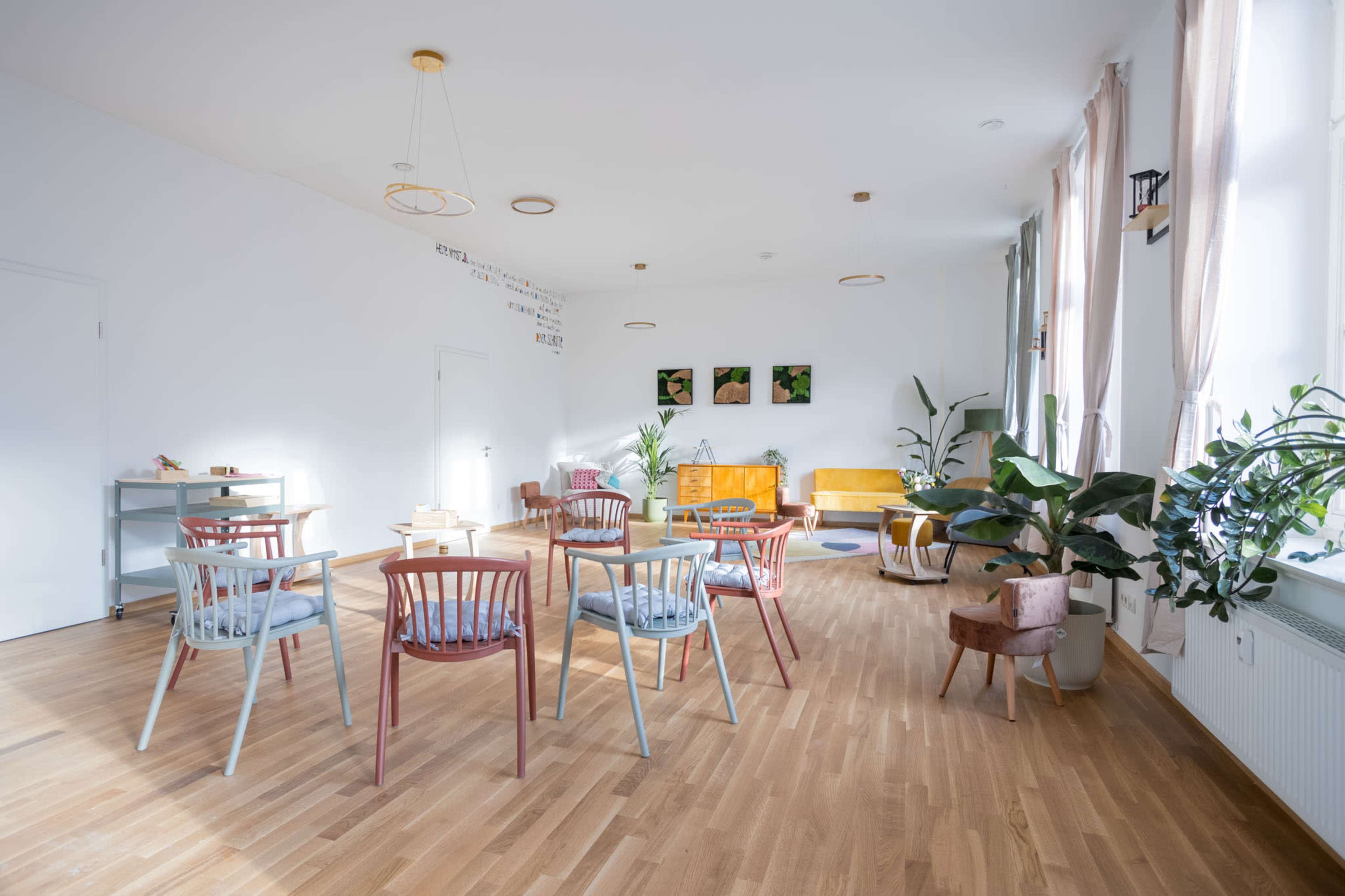 The image shows a bright, modern room with wooden flooring, featuring several colorful chairs arranged around small tables, and potted plants placed around the space.