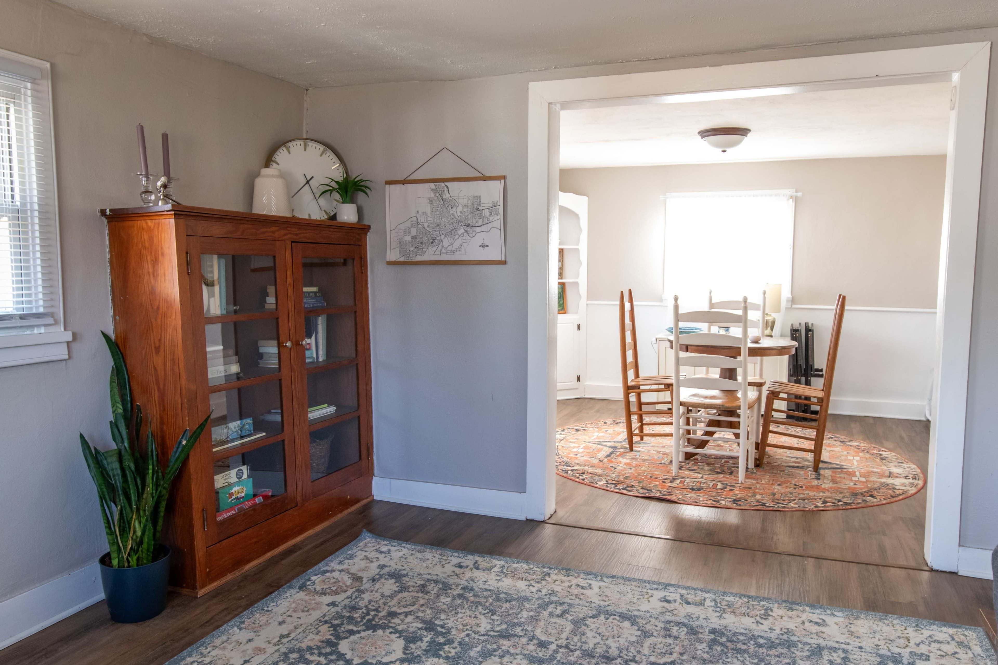 The image shows a bright interior space with a wooden cabinet on the left, a plant in a pot, and a dining area featuring a round table and chairs in the adjacent room.