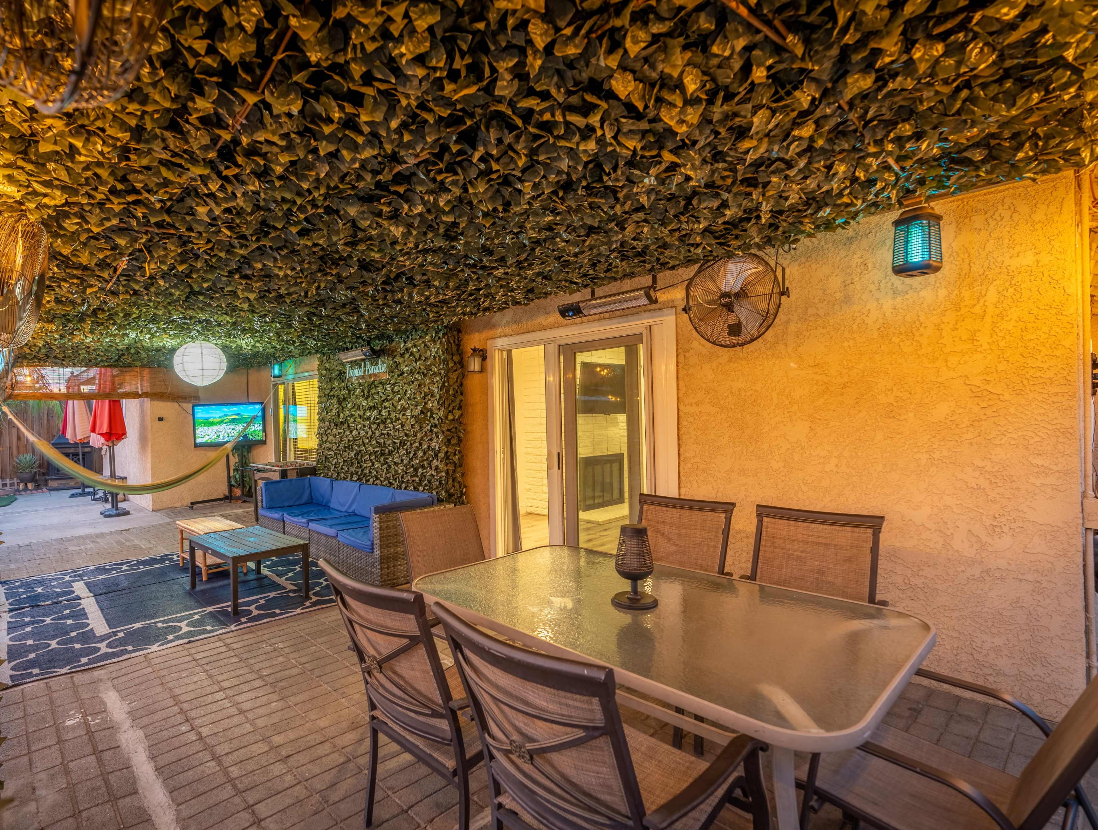 A covered outdoor patio with a seating area, table, and ceiling adorned with green foliage.