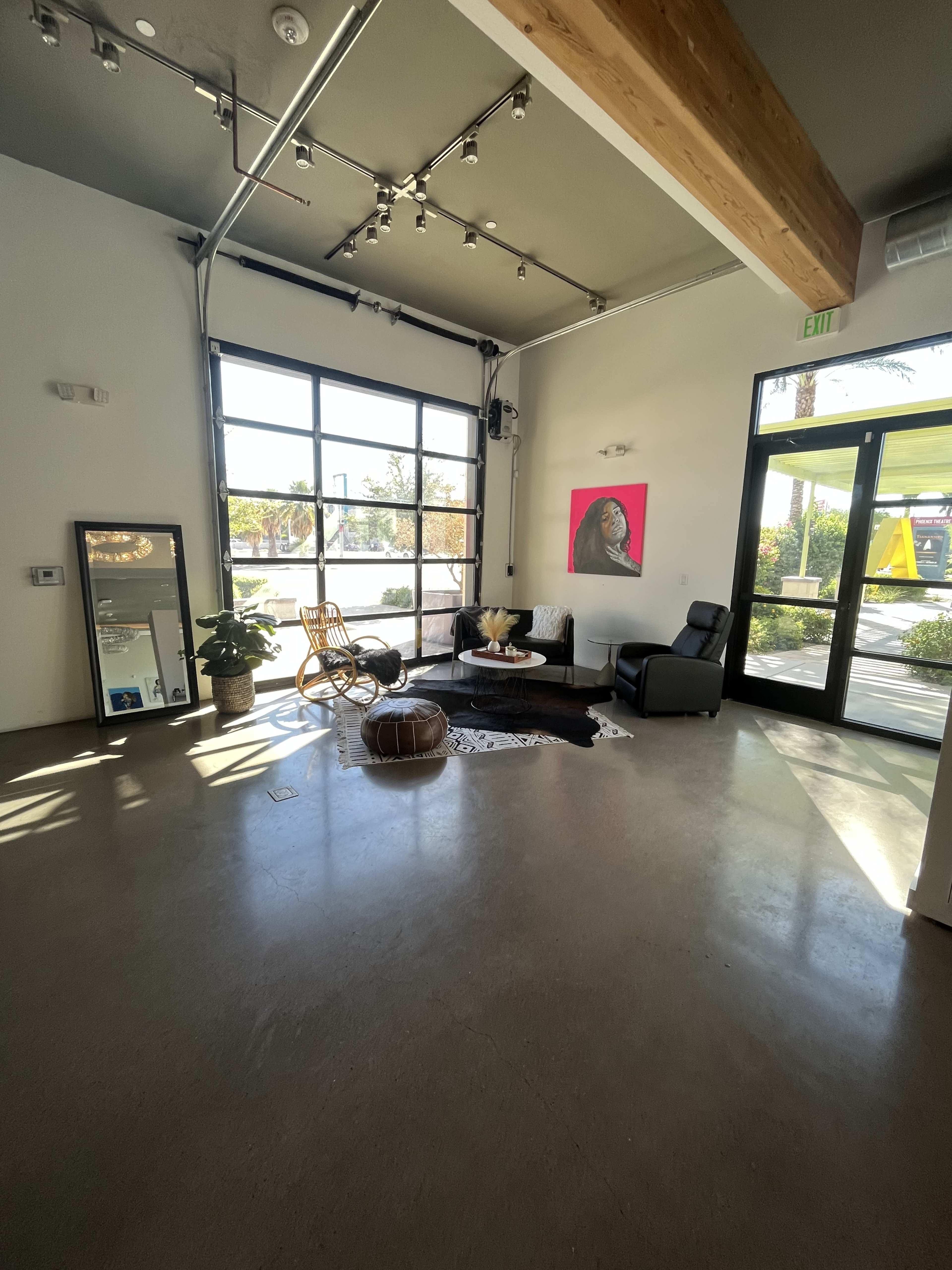 The image shows a spacious room with large windows, featuring a modern seating area with a chair, a small table, and a mirror against the wall.