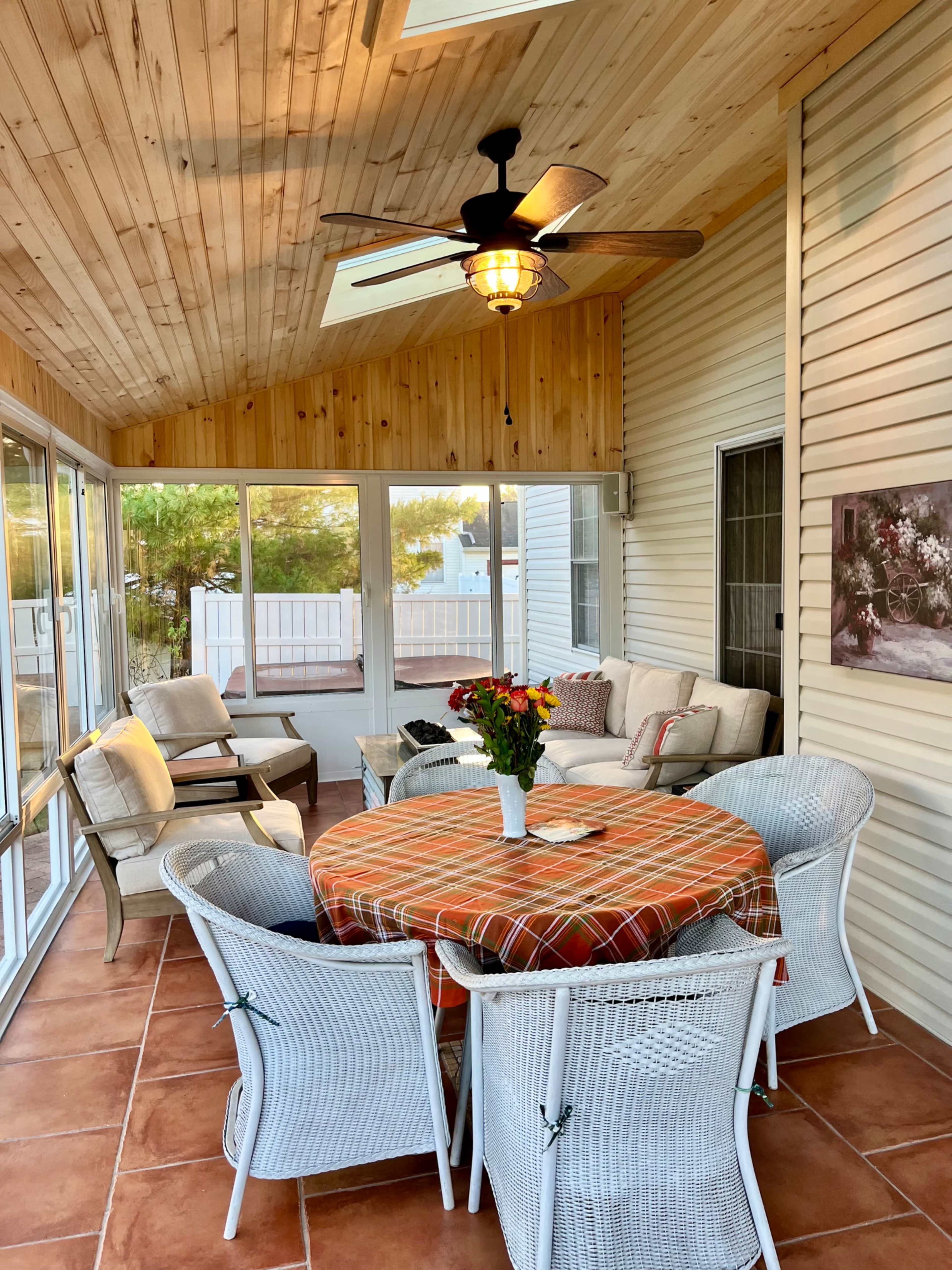 The image shows a sunroom with wooden ceilings, a round table covered by a plaid tablecloth, four white woven chairs, and a seating area with a sofa and armchairs near a window.