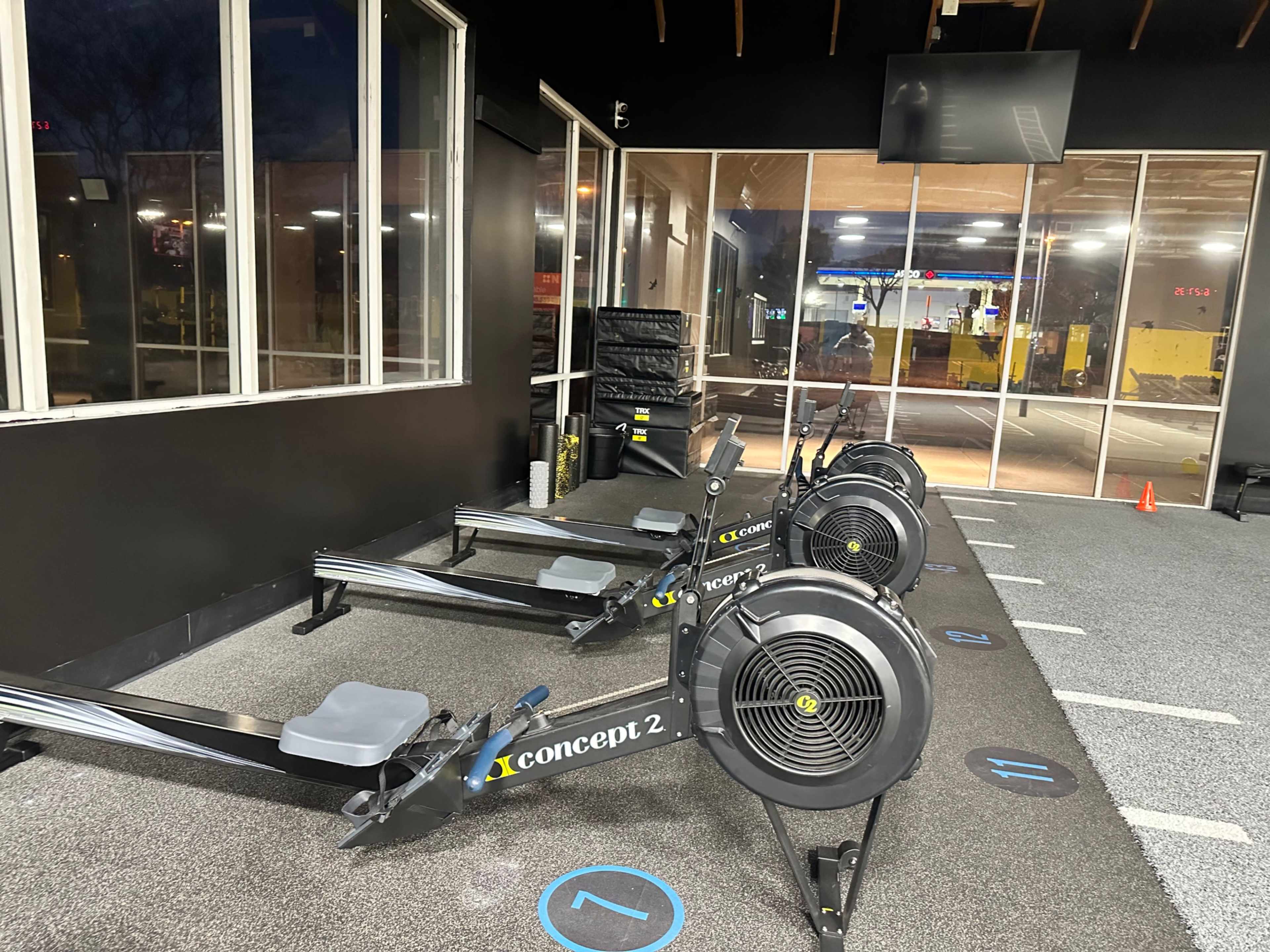 Several Concept 2 rowing machines lined up in a gym, with large windows displaying a night view outside.