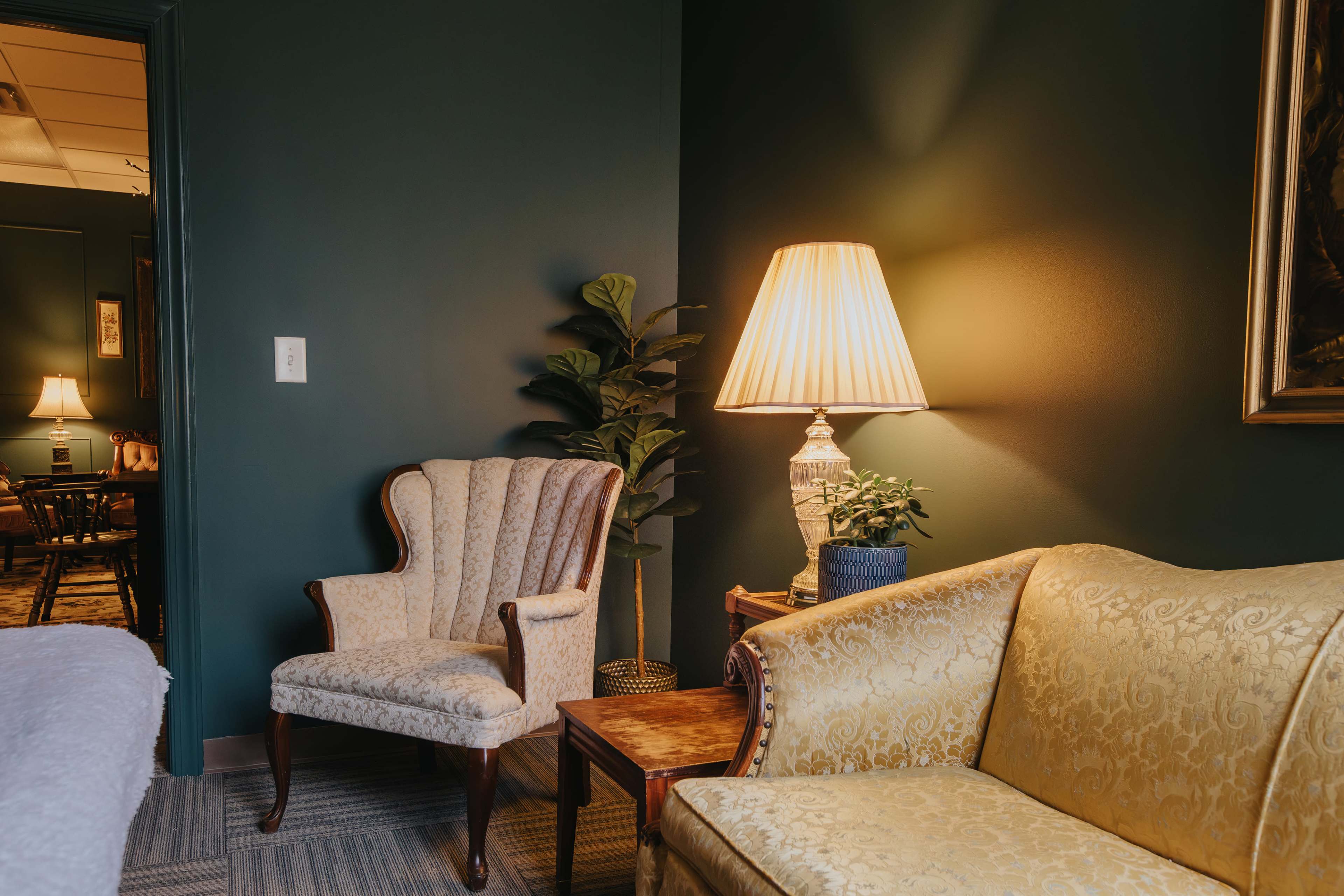 The scene features a cozy room with a yellow sofa, an antique armchair, a table lamp, and a potted plant against a dark green wall.