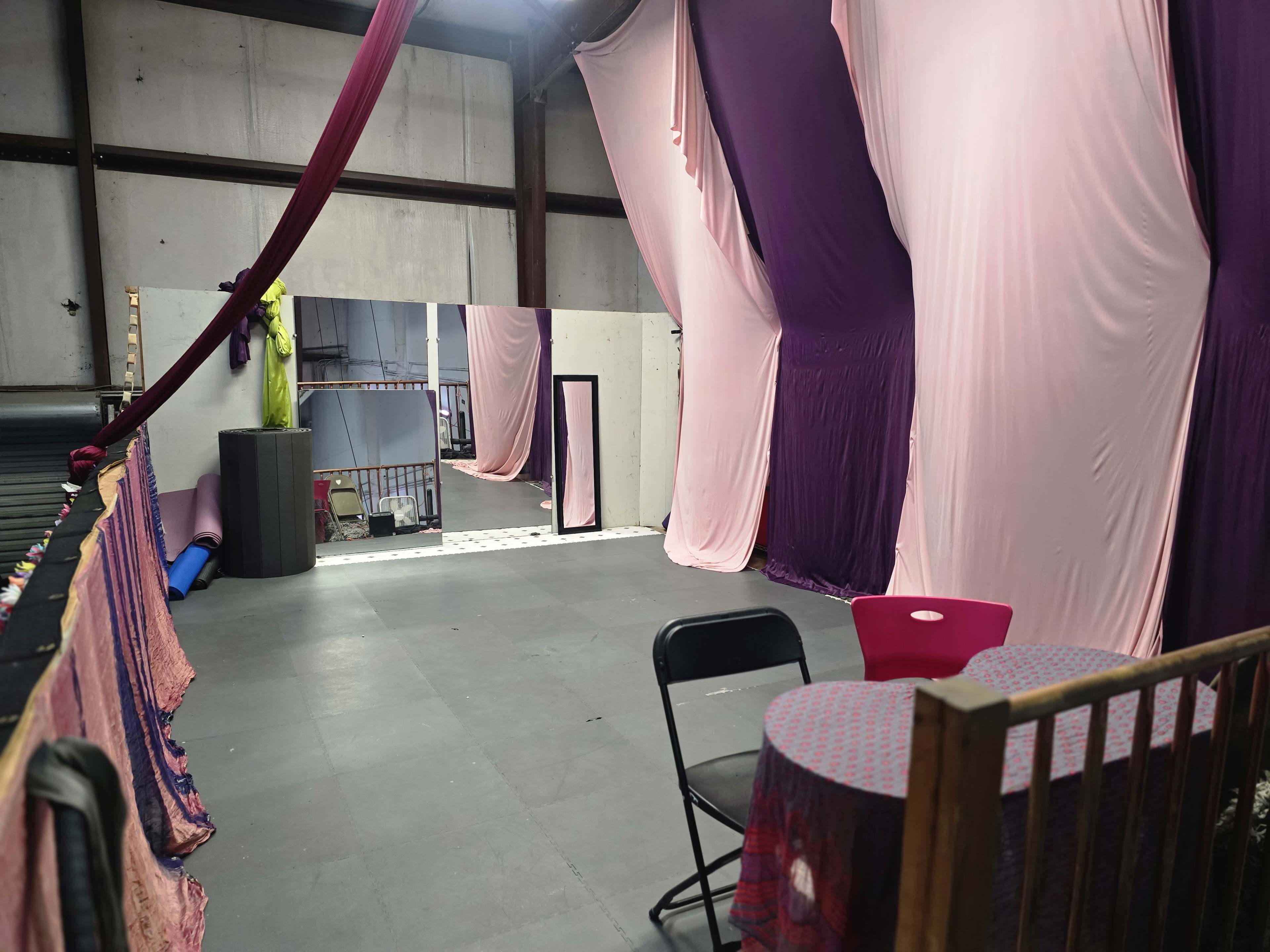 An indoor space with draped fabric in pink and purple, a mirror, a small round table with chairs, and a statue on a pedestal in the background.
